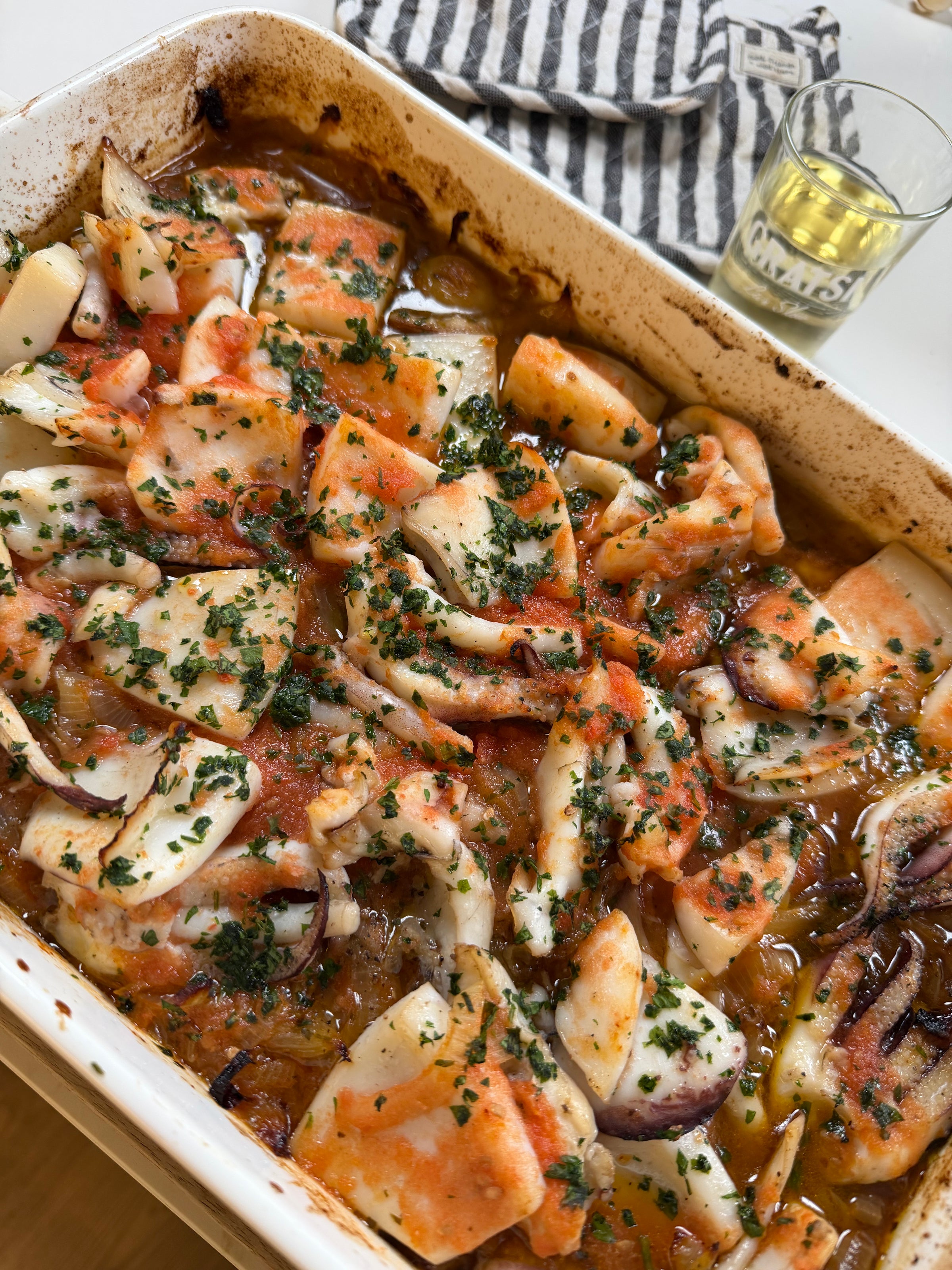 A baked seafood dish with pieces of squid in a tomato-based sauce, garnished with chopped herbs, served in a white rectangular dish. A glass of white wine and a striped napkin are in the background.