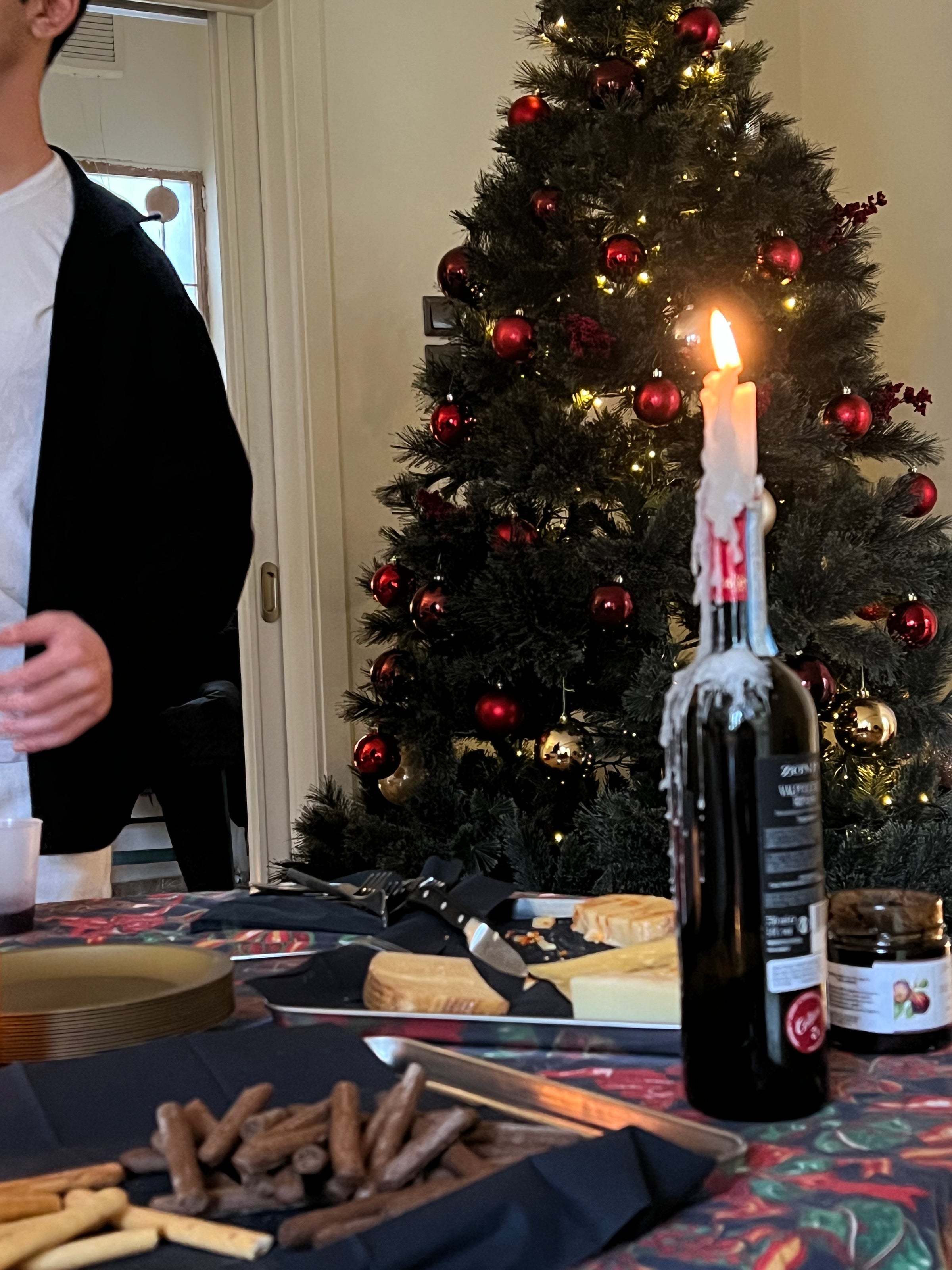 A Christmas tree with red and gold ornaments stands behind a table set with snacks, cheese, and a wine bottle holding a lit candle. A person in a white shirt and black sweater is partially visible on the left.