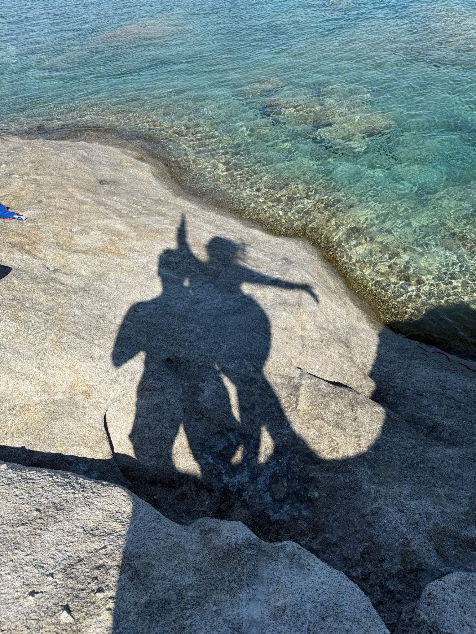 Two people’s shadows are cast on a rocky surface near clear, blue-green water. One person appears to be pointing or raising their arm while both are standing close together.