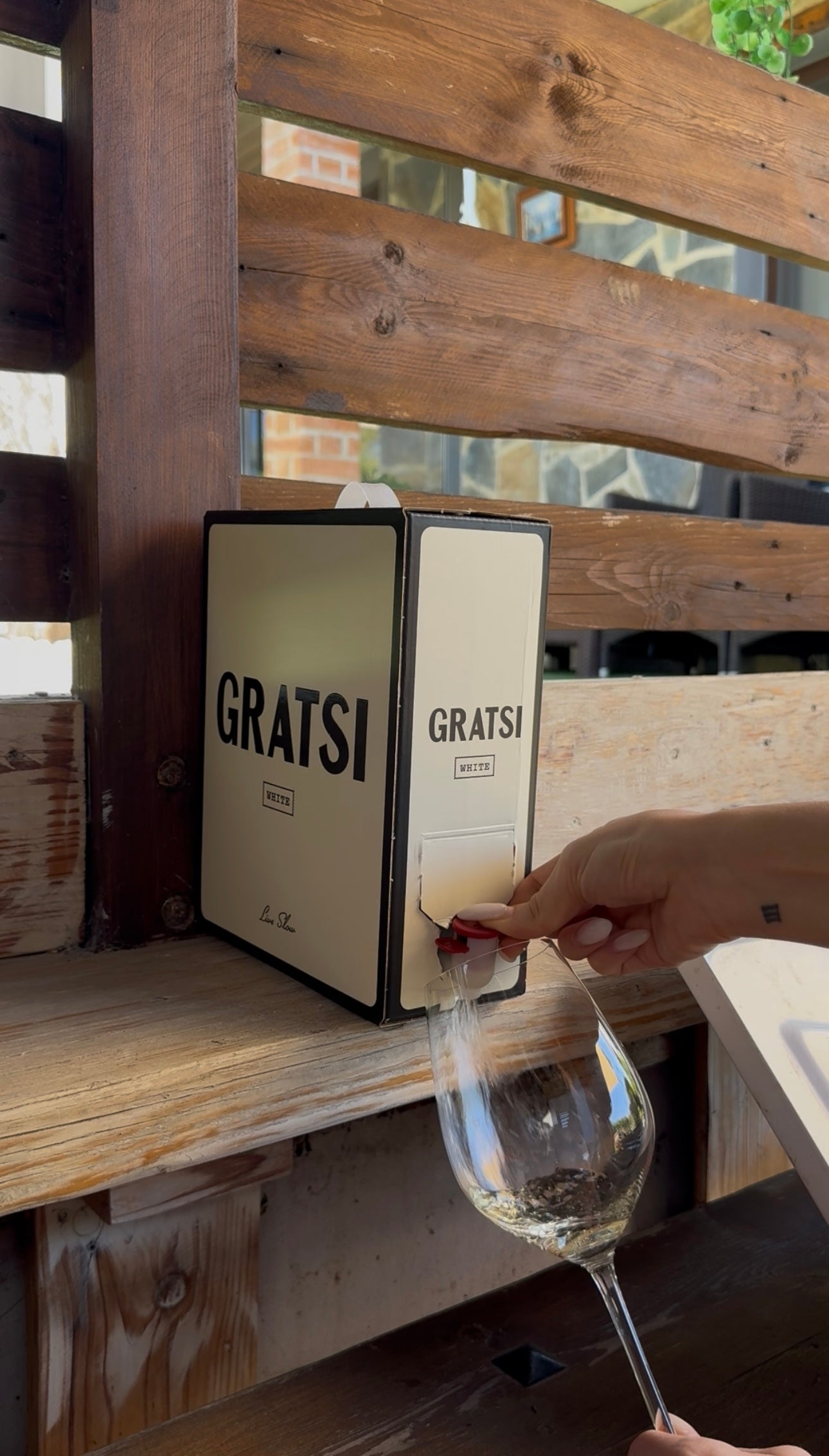 A hand dispenses white wine from a Gratsi box into a clear wine glass on a wooden outdoor counter, with sunlight streaming through wooden slats in the background.