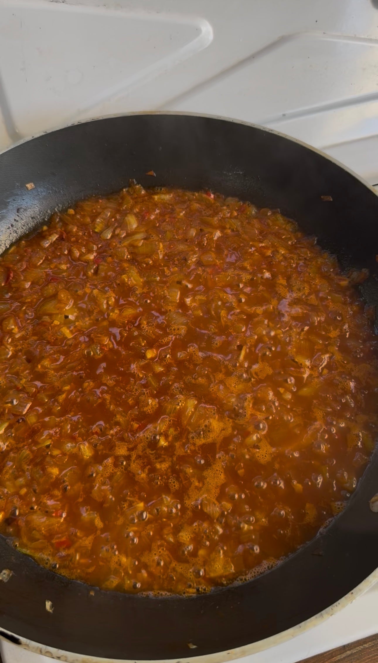 A close-up of a pan on a stovetop filled with simmering red-orange sauce containing chopped onions and spices, likely a tomato-based mixture, with steam rising gently.