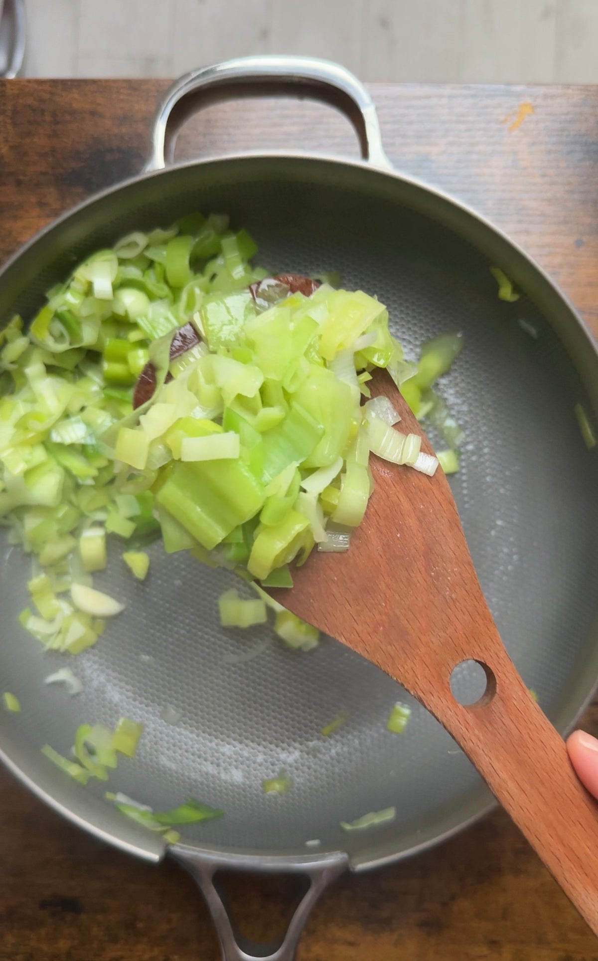A wooden spoon lifts chopped leeks from a frying pan.