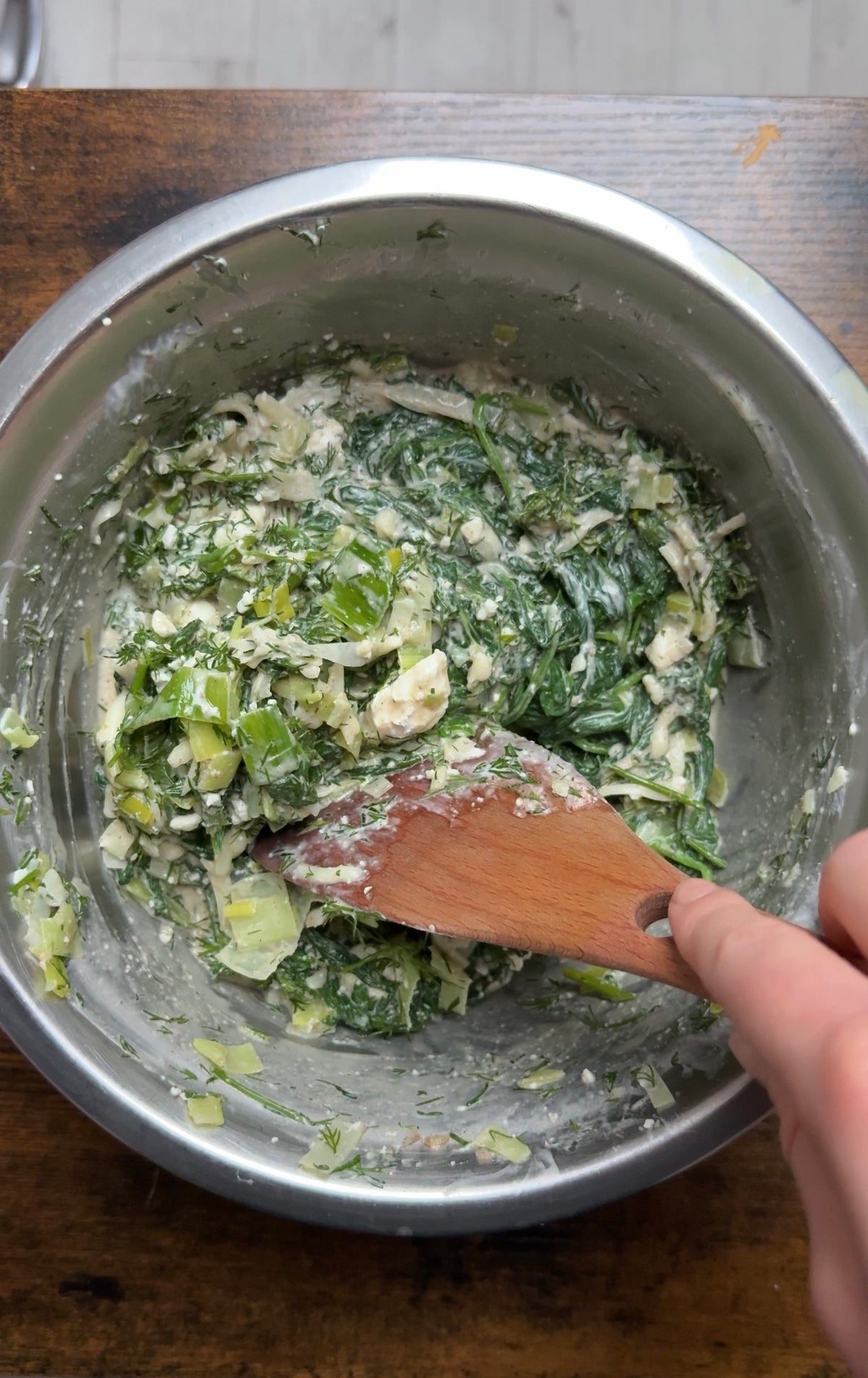 A hand uses a wooden spoon to stir a mixture of chopped greens, fresh dill, and crumbled cheese in a metal mixing bowl.