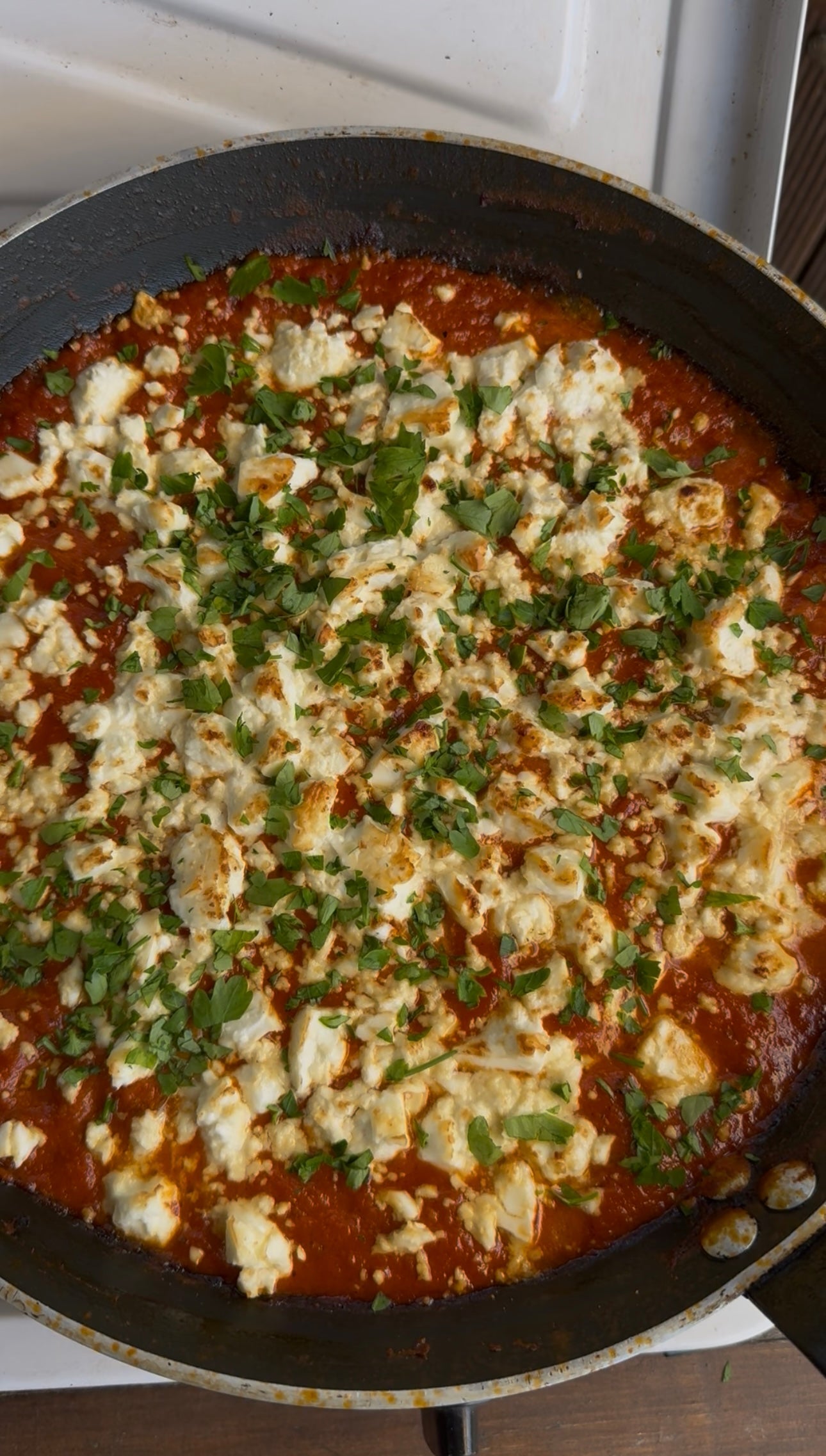A close-up of a pan filled with a tomato-based dish, topped with crumbled white cheese and garnished with chopped fresh herbs, likely cilantro or parsley. The dish is on a stovetop.