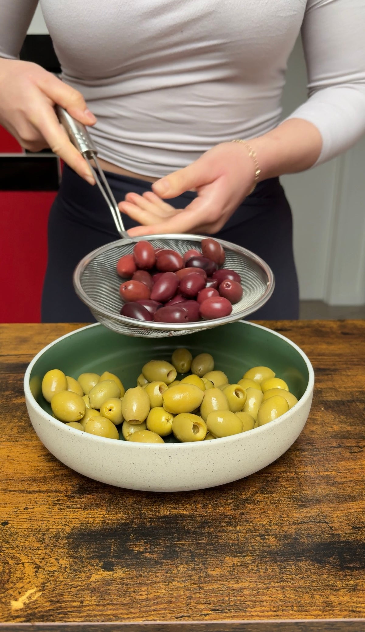 A person holding a metal strainer full of dark purple olives over a wide bowl filled with green olives.
