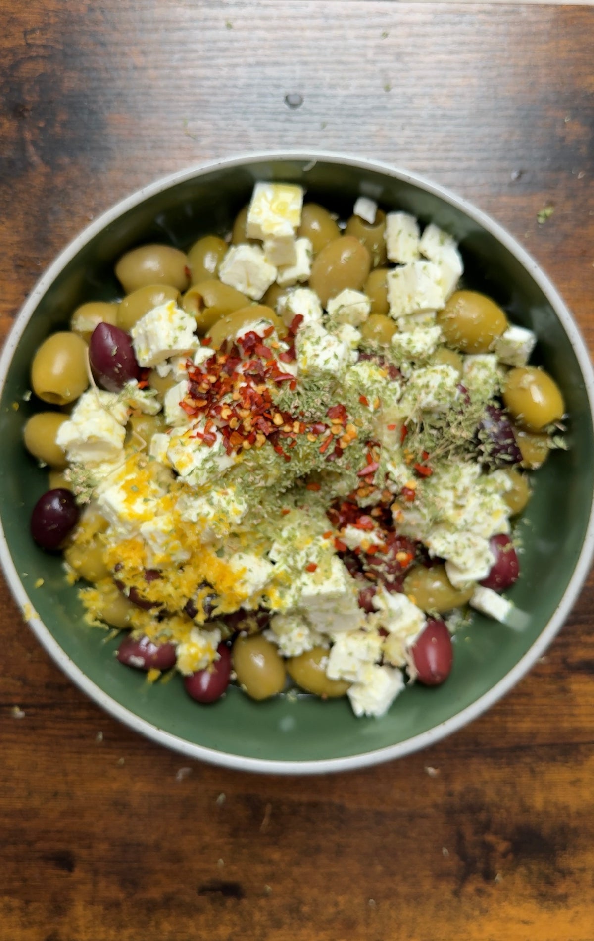 A bowl of green and black olives topped with cubes of feta cheese, dried herbs, and red pepper flakes.