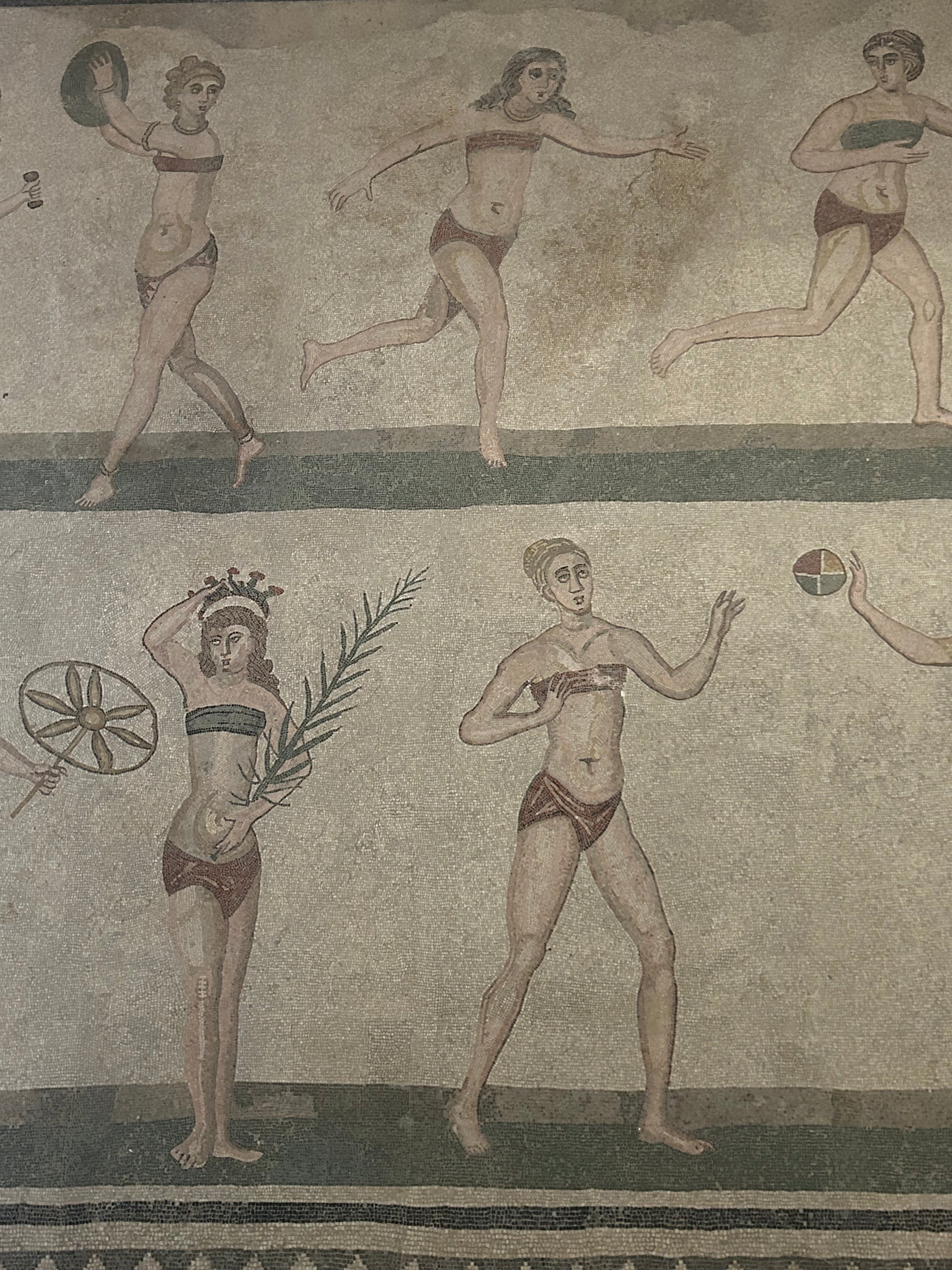 Ancient Roman mosaic showing women in bikinis playing sports, holding a discus, ball, and palm branch, set against a neutral background.