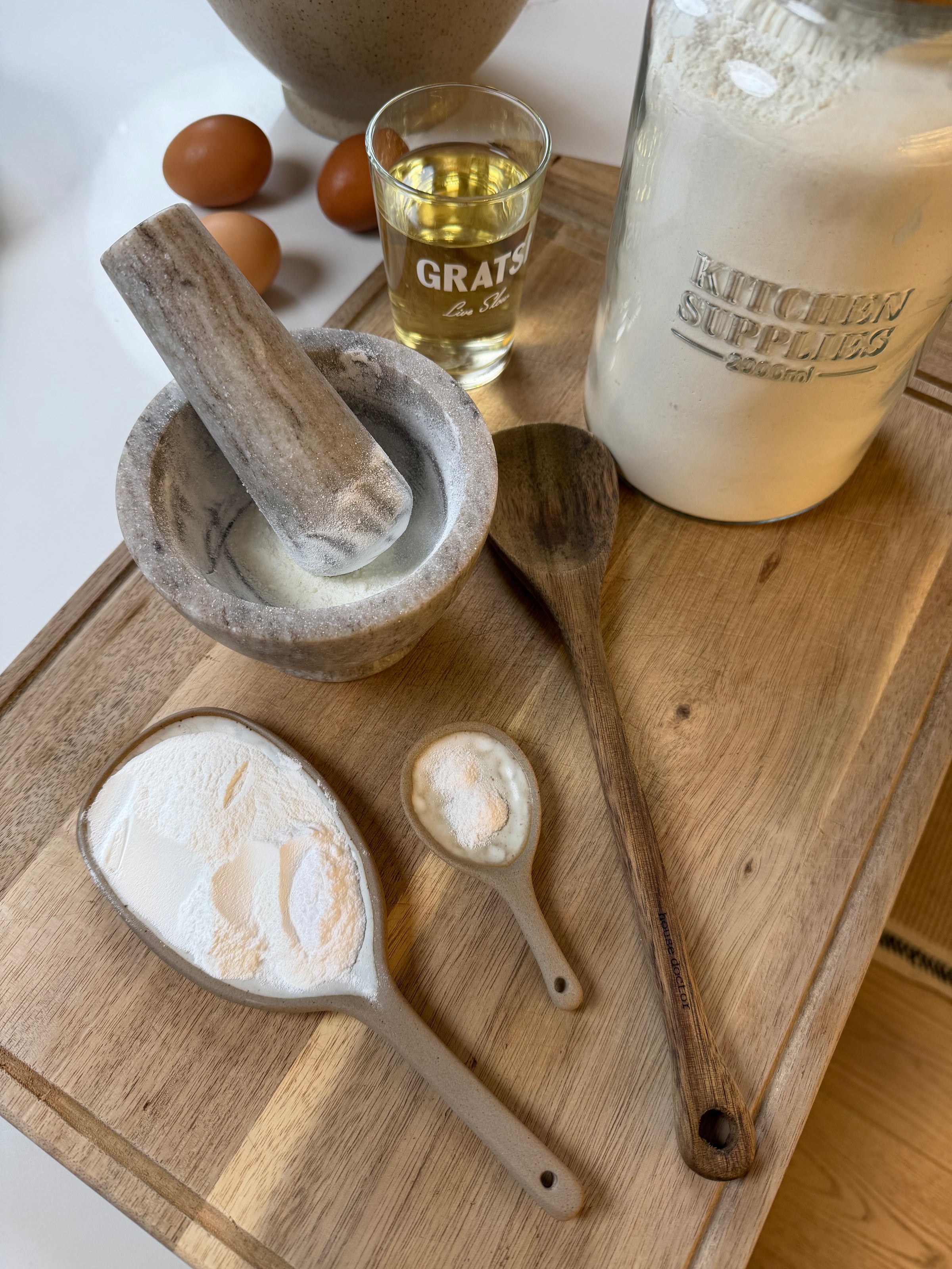 A wooden board with a jar of flour, two ceramic measuring spoons with flour and baking powder, a wooden spoon, a small stone mortar and pestle, a glass of oil, and three eggs in the background.