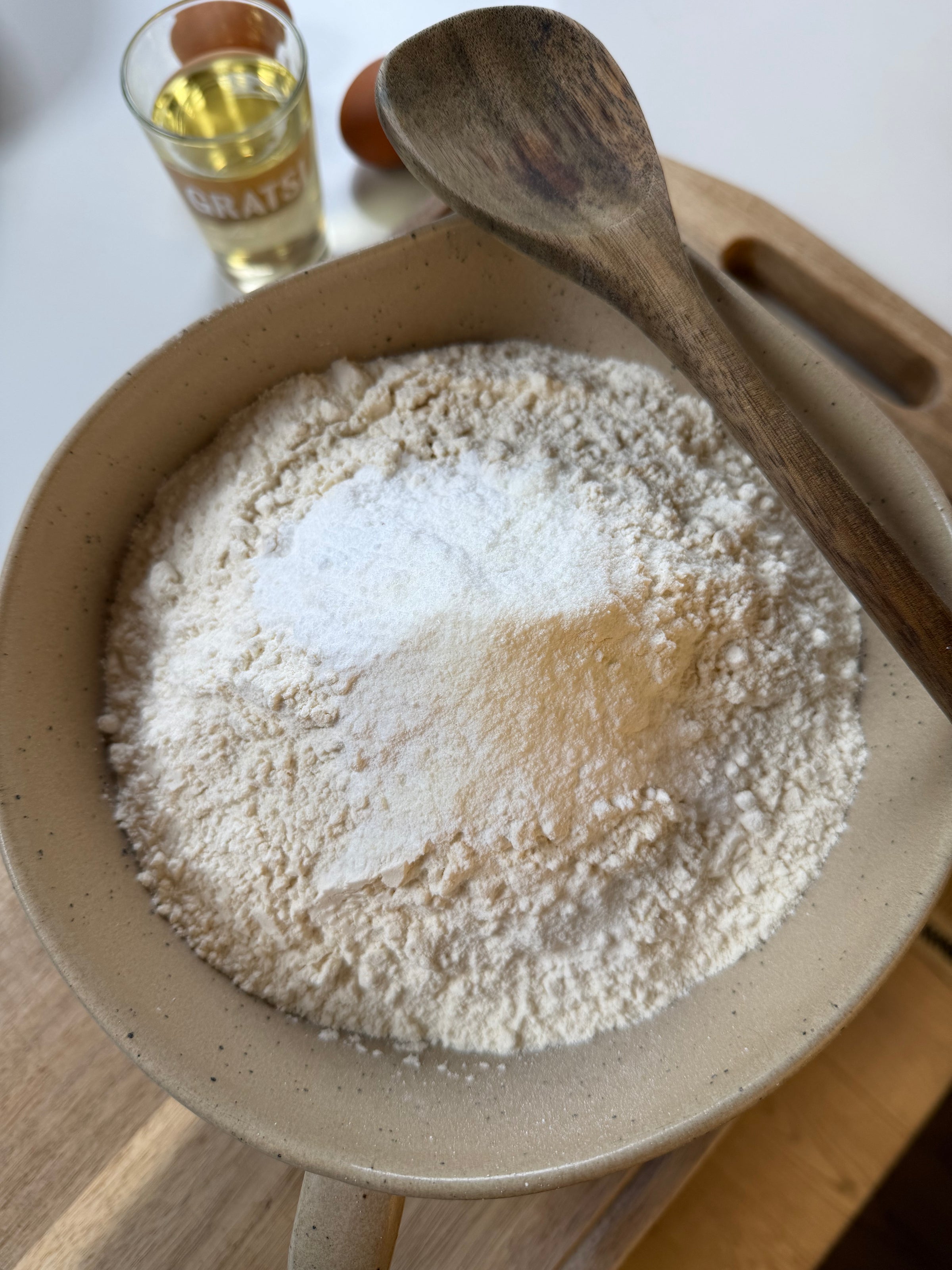 A beige bowl filled with flour and baking powder sits on a wooden board, with a wooden spoon resting on top. In the background, a glass of liquid and two eggs are partially visible.