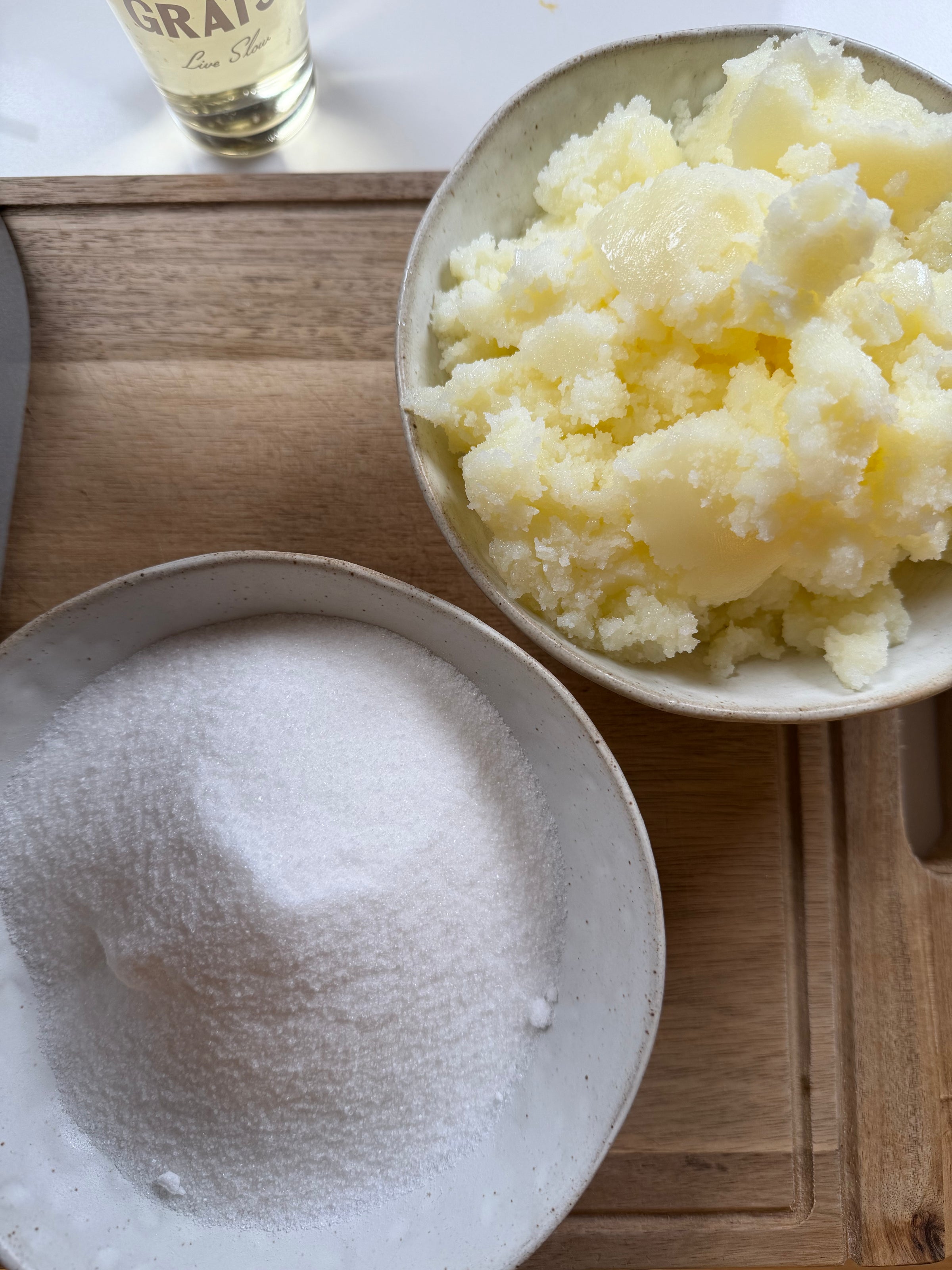 Two white bowls on a wooden tray: one filled with granulated white sugar, the other with chunks of pale yellow butter. A small part of a glass bottle is visible in the top corner.