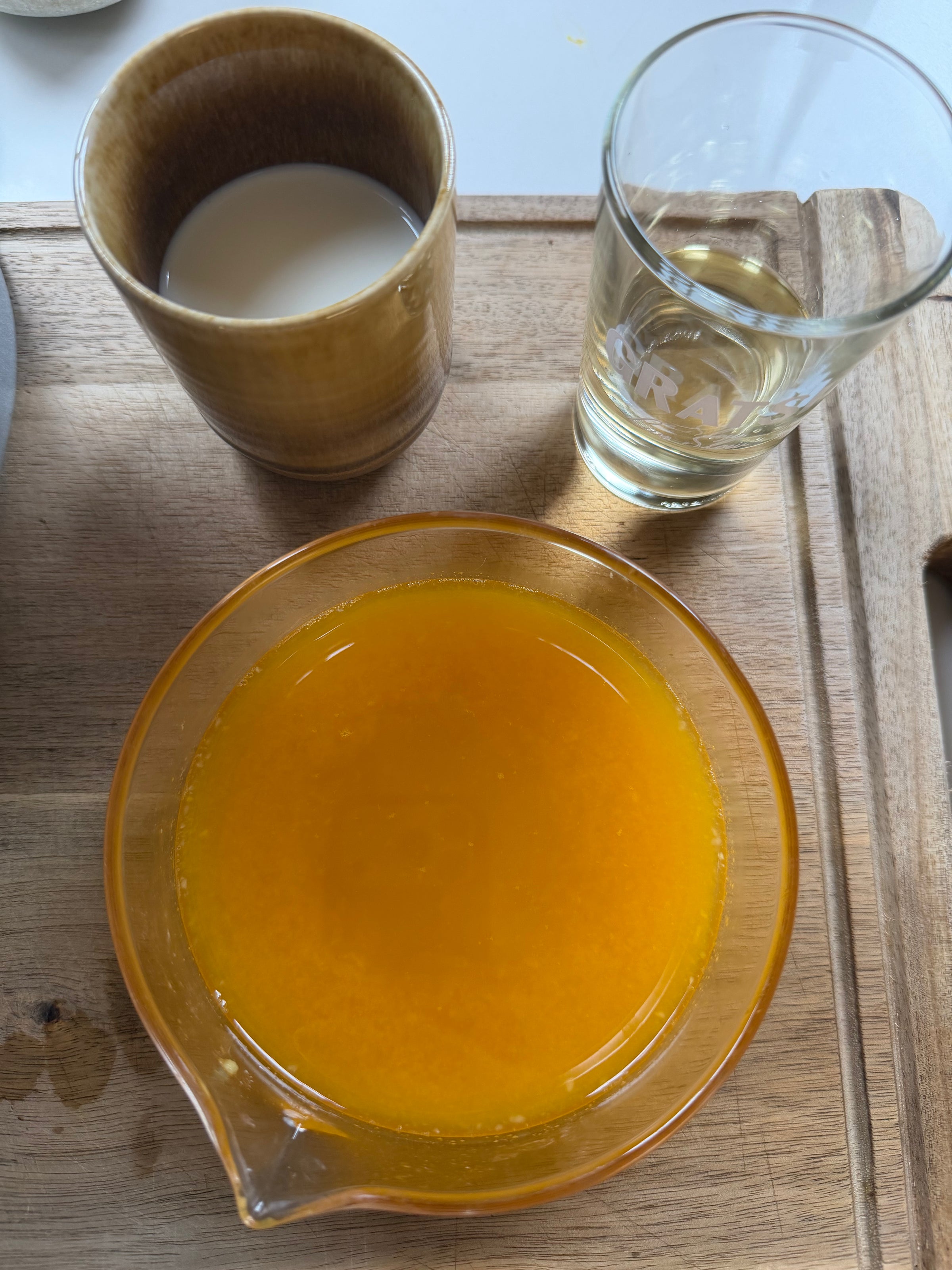 A wooden tray holds a cup of milk tea, a glass of clear liquid, and a bowl of bright orange liquid, possibly melted butter, on a wooden surface.