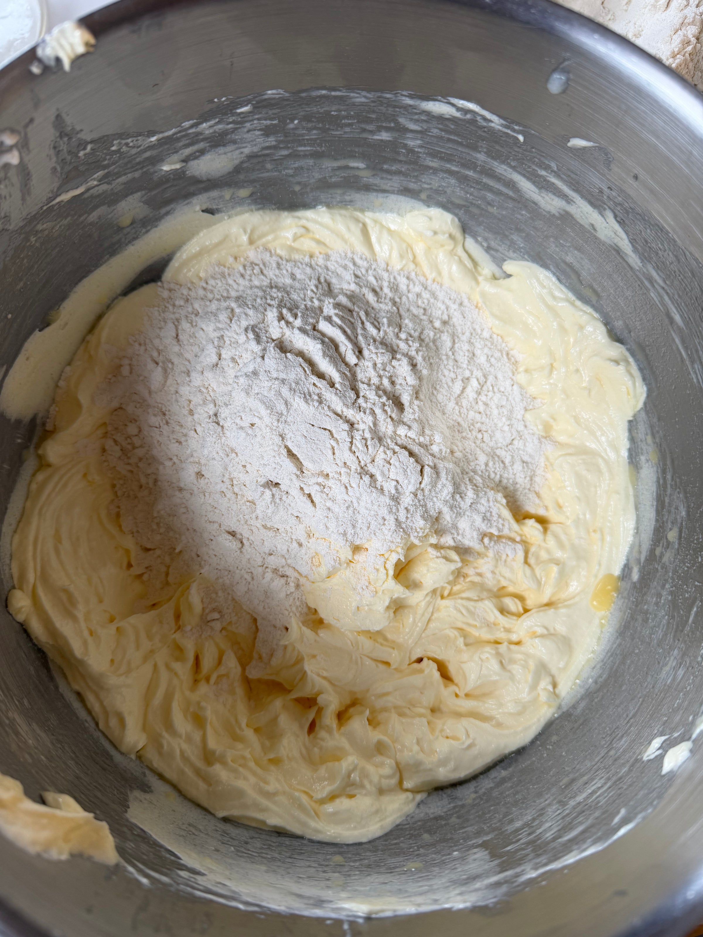 A metal mixing bowl containing creamy yellow batter with a mound of flour on top, ready to be mixed in.