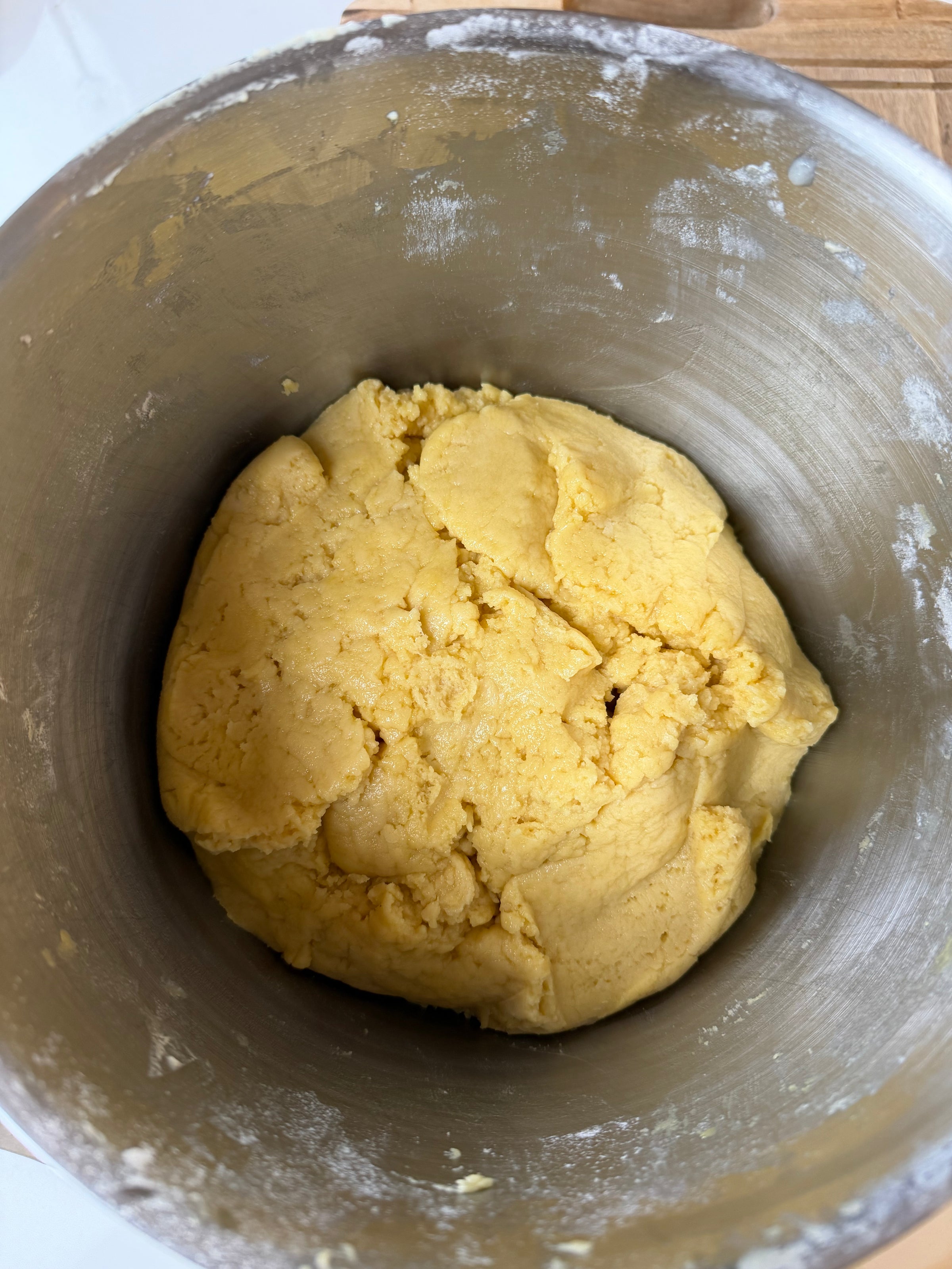 A ball of yellow dough sits in the bottom of a stainless steel mixing bowl, with bits of flour dusted along the sides.
