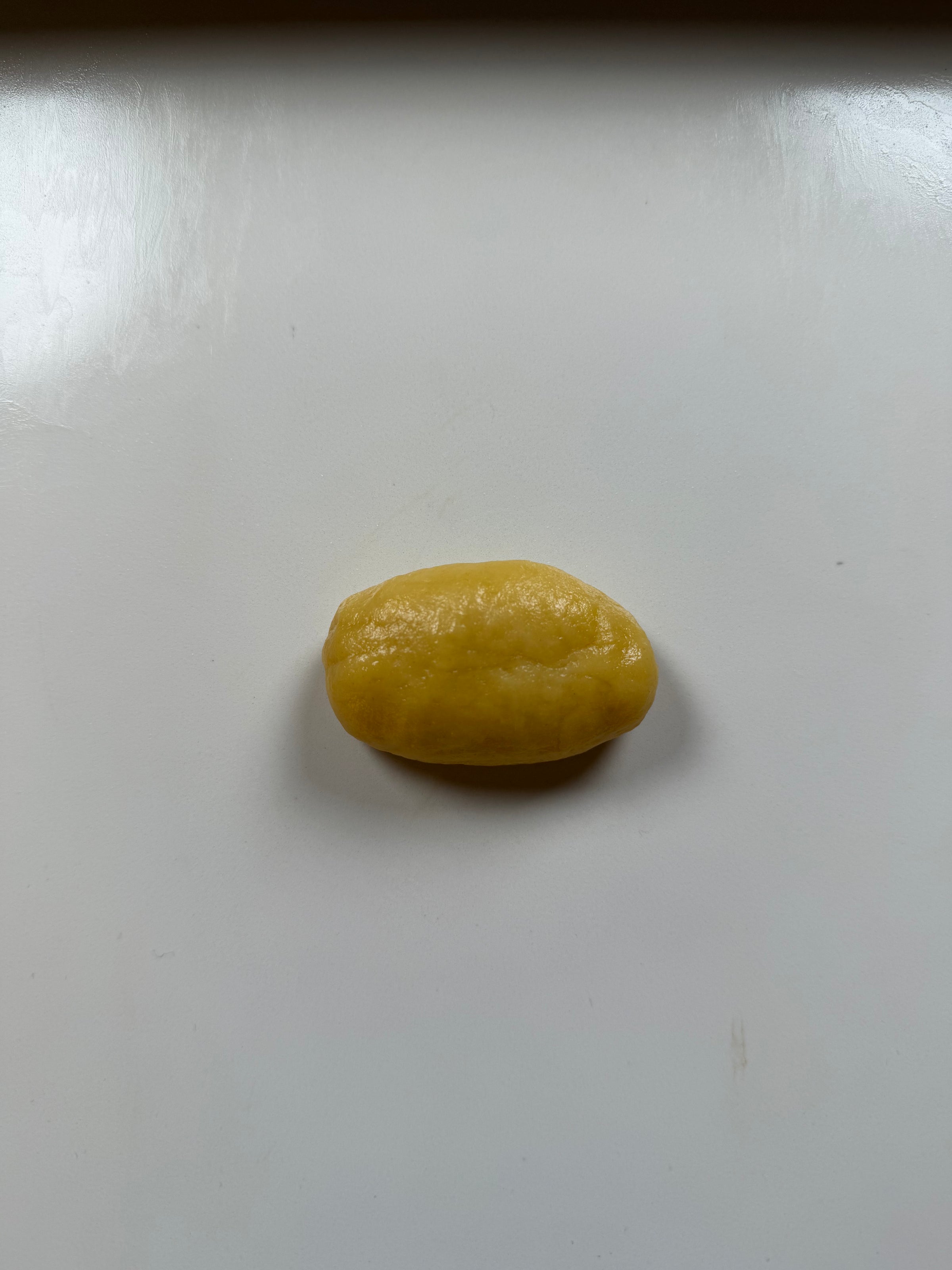 A small, oval-shaped ball of yellow dough sits on a smooth, white surface under soft lighting.