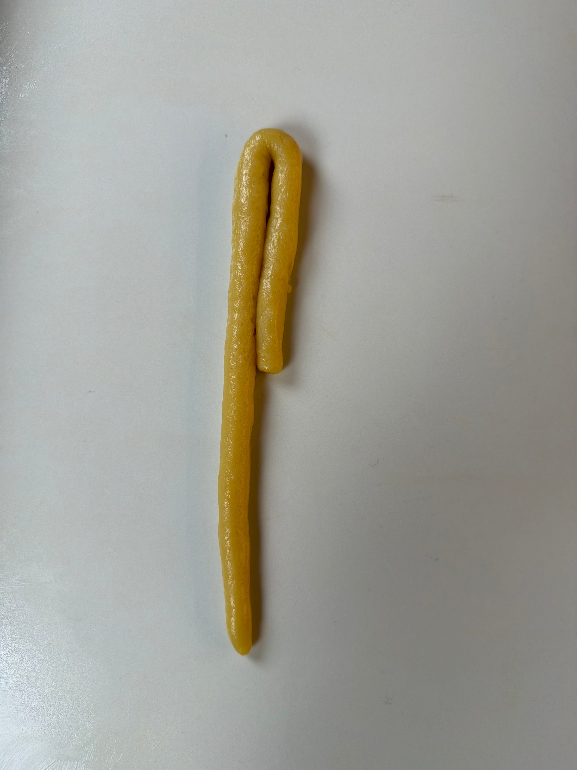 A single, yellow beeswax candle shaped like a shepherds crook, lying flat on a plain, light gray surface.