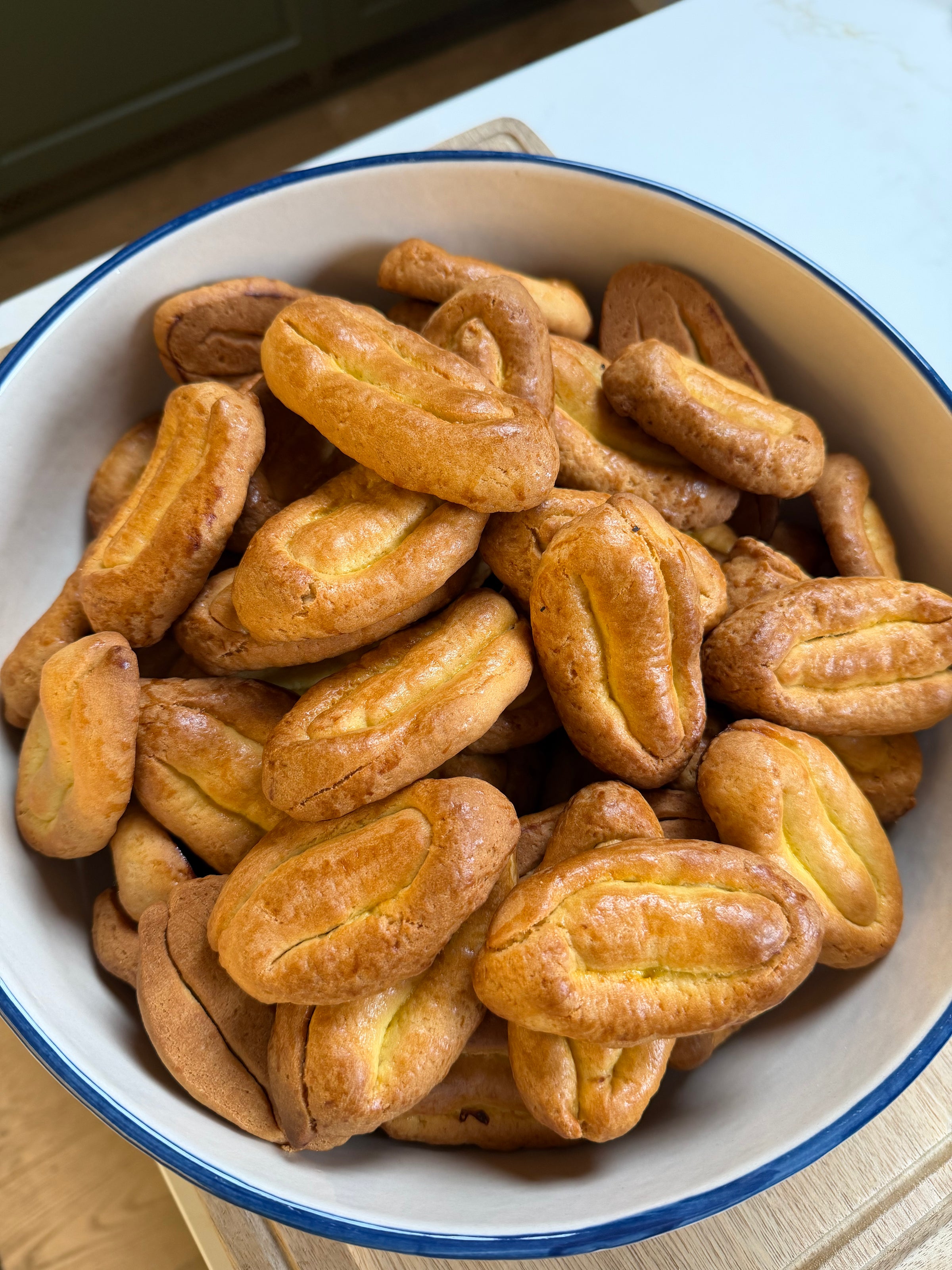 A white bowl with a blue rim filled with oval-shaped, golden-brown baked cookies, all arranged closely together. The cookies have a slightly shiny surface and a central indentation.