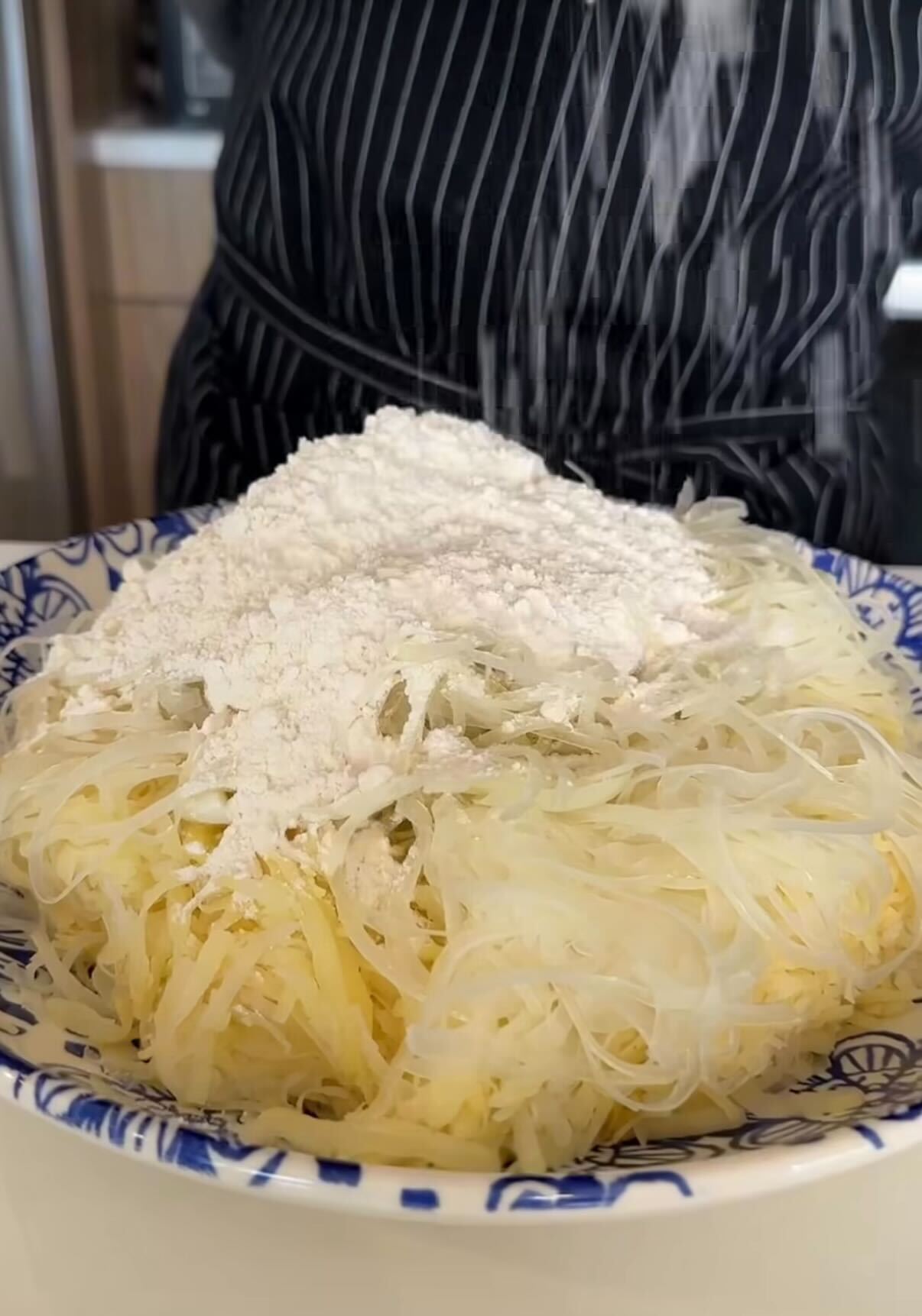 A bowl filled with shredded potatoes and a mound of flour, with a person in a striped apron visible in the background, sprinkling more flour on top.