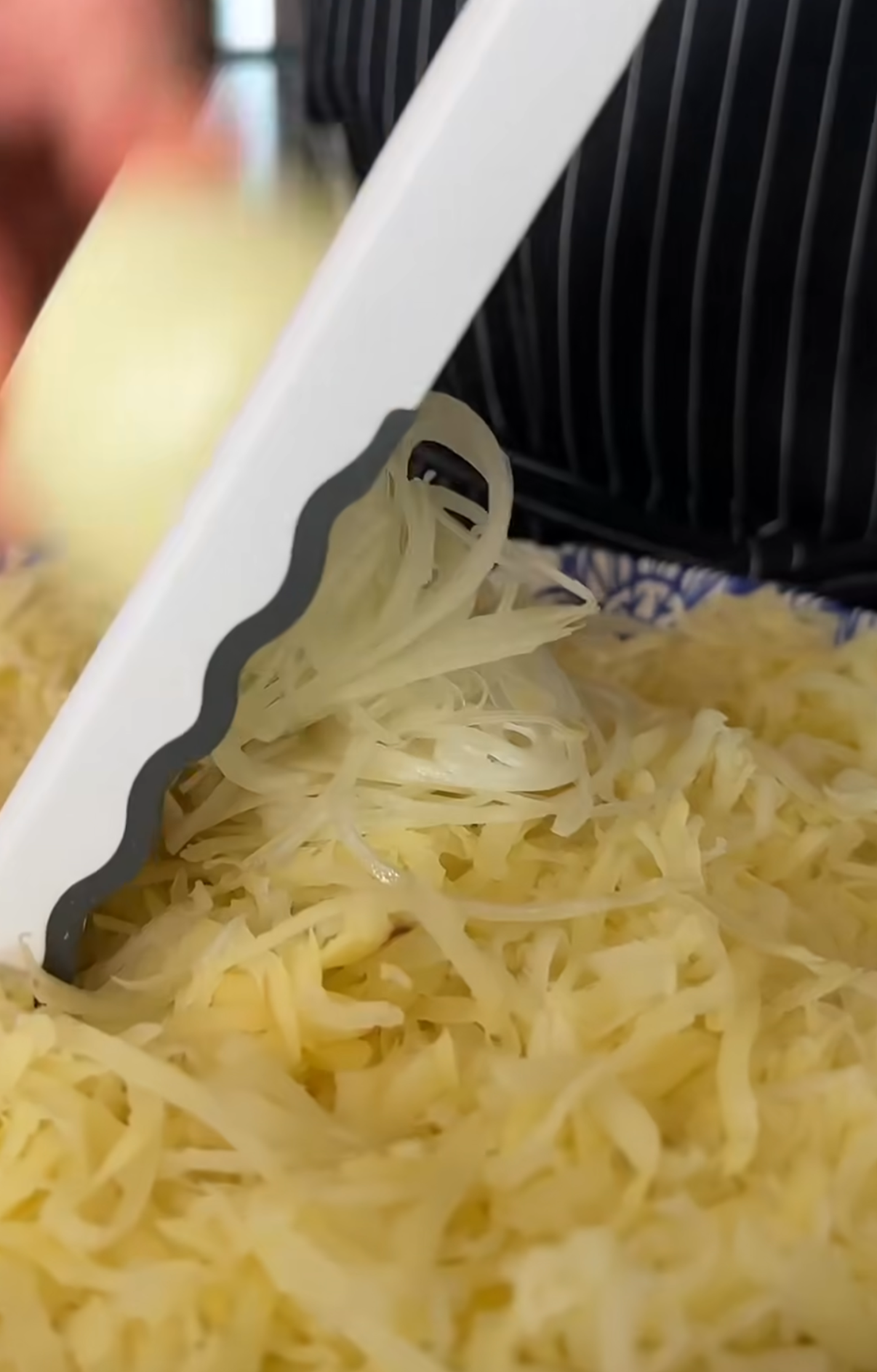 A close-up of a hand slicing a potato with a mandoline slicer, creating thin potato strips that pile up below. The person is wearing a black and white striped apron.