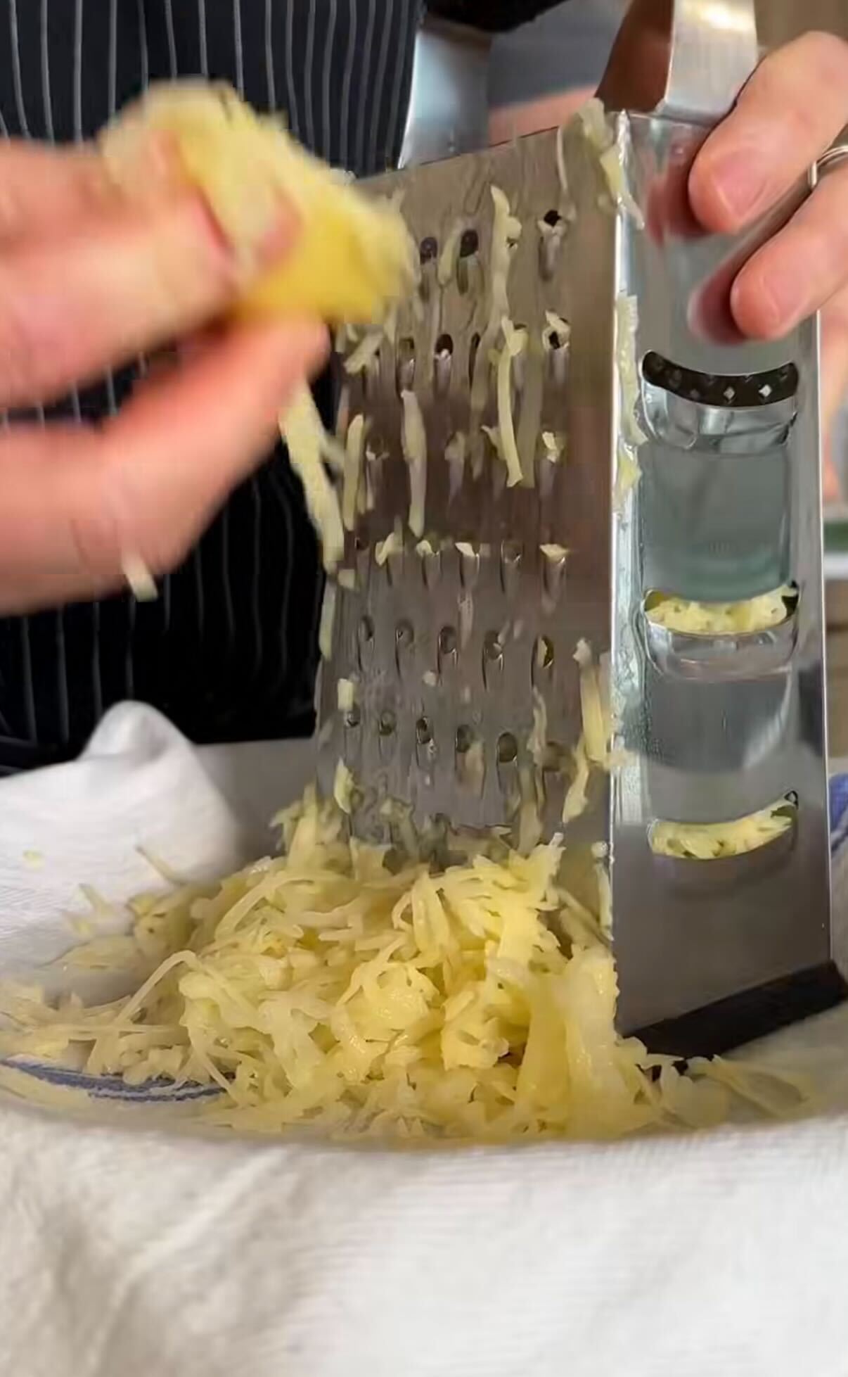A person grates a block of cheese using a metal box grater, with shredded cheese collecting on a white cloth below.