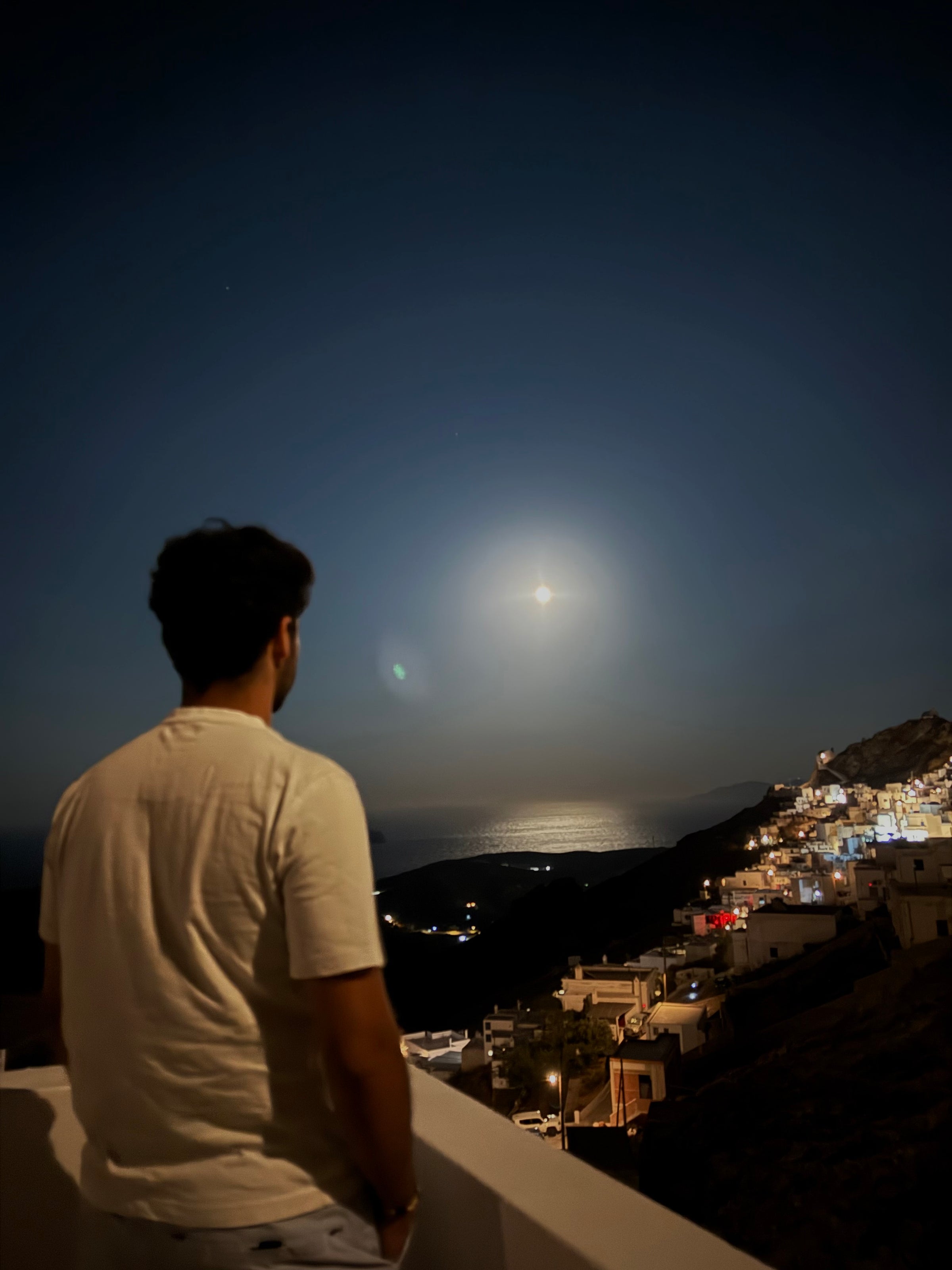 A person stands on a balcony at night, looking at the full moon over a coastal town with illuminated buildings and the sea reflecting the moonlight.
