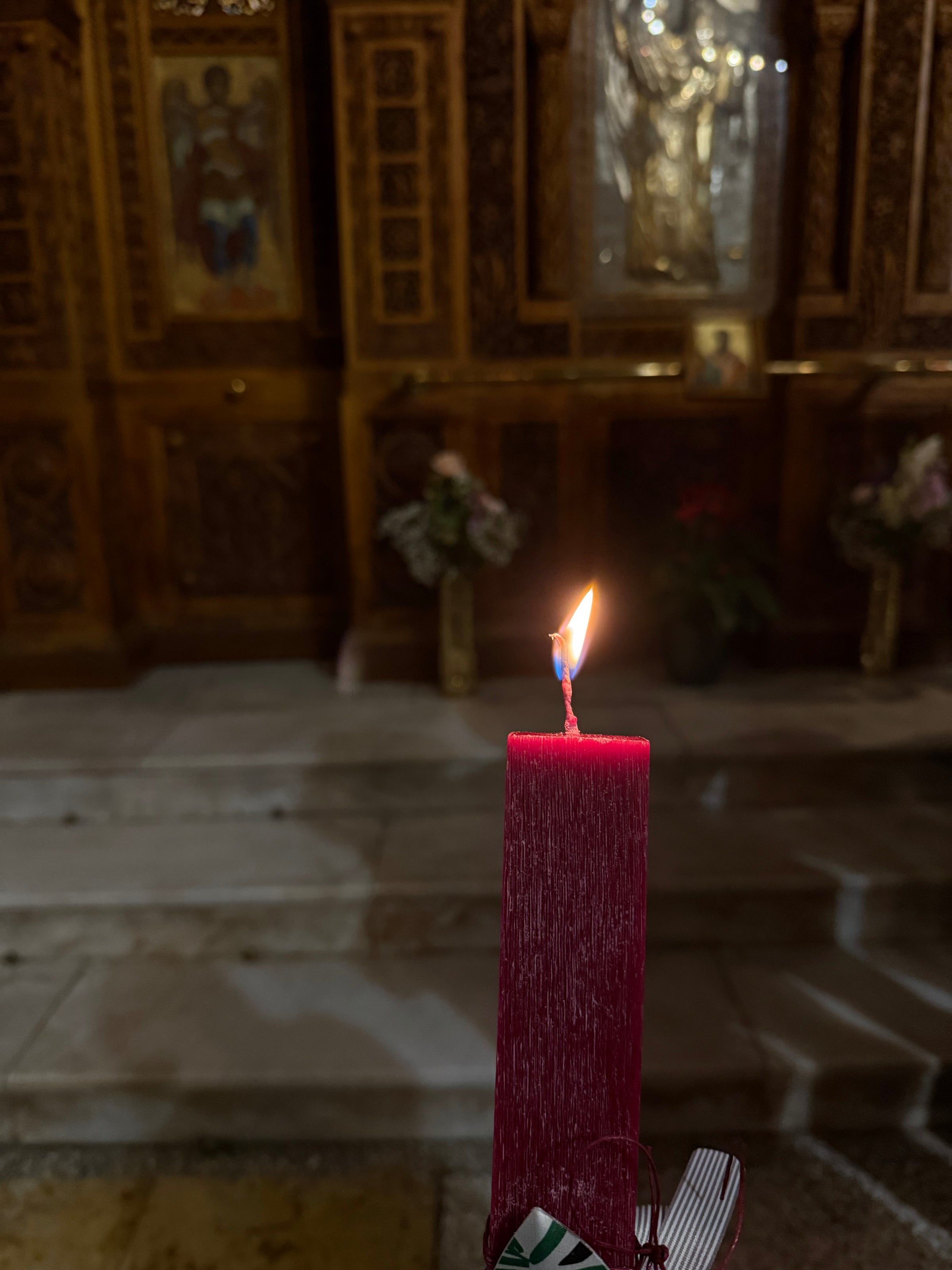 A red candle with a lit flame is held in the foreground inside a dimly lit church, with ornate wooden panels, icons, and flower arrangements visible in the background.