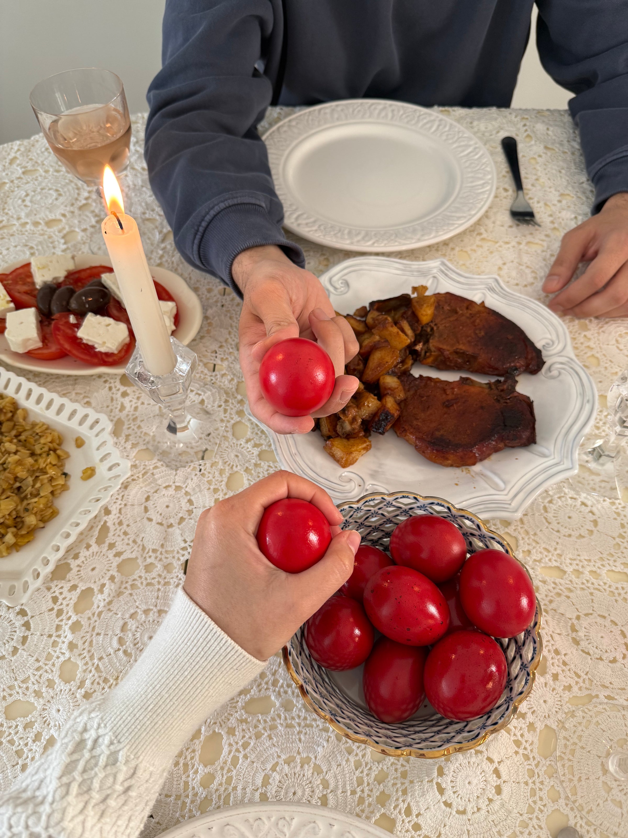 Two people at a table with ornate plates and food tap red eggs together, a lit candle nearby. The tablecloth is lace, and a bowl of red eggs is in the foreground.