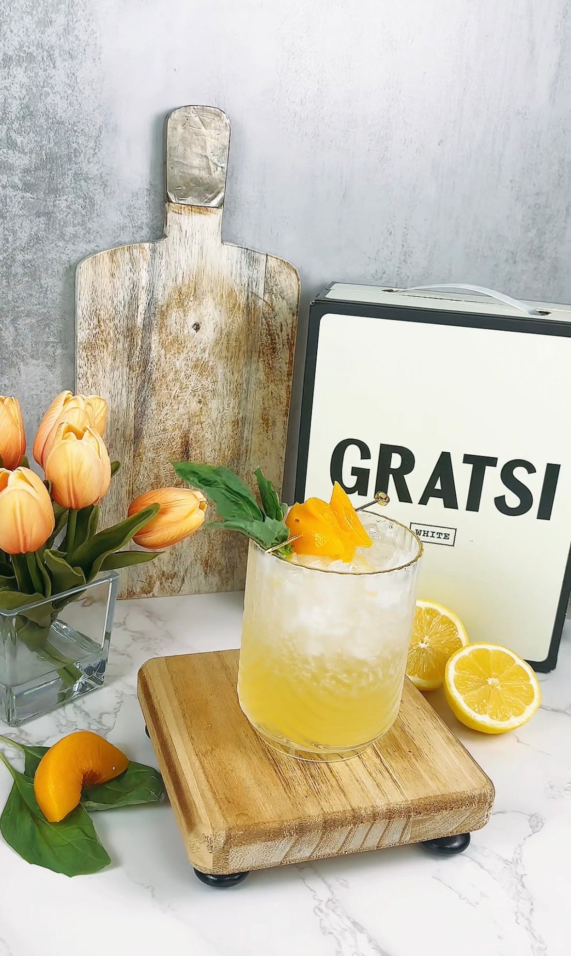 A yellow cocktail with ice, garnished with an orange slice and mint, sits on a wooden board. Nearby are halved lemons, a peach slice, orange tulips in a vase, a rustic cutting board, and a box labeled “GRATSI.”.