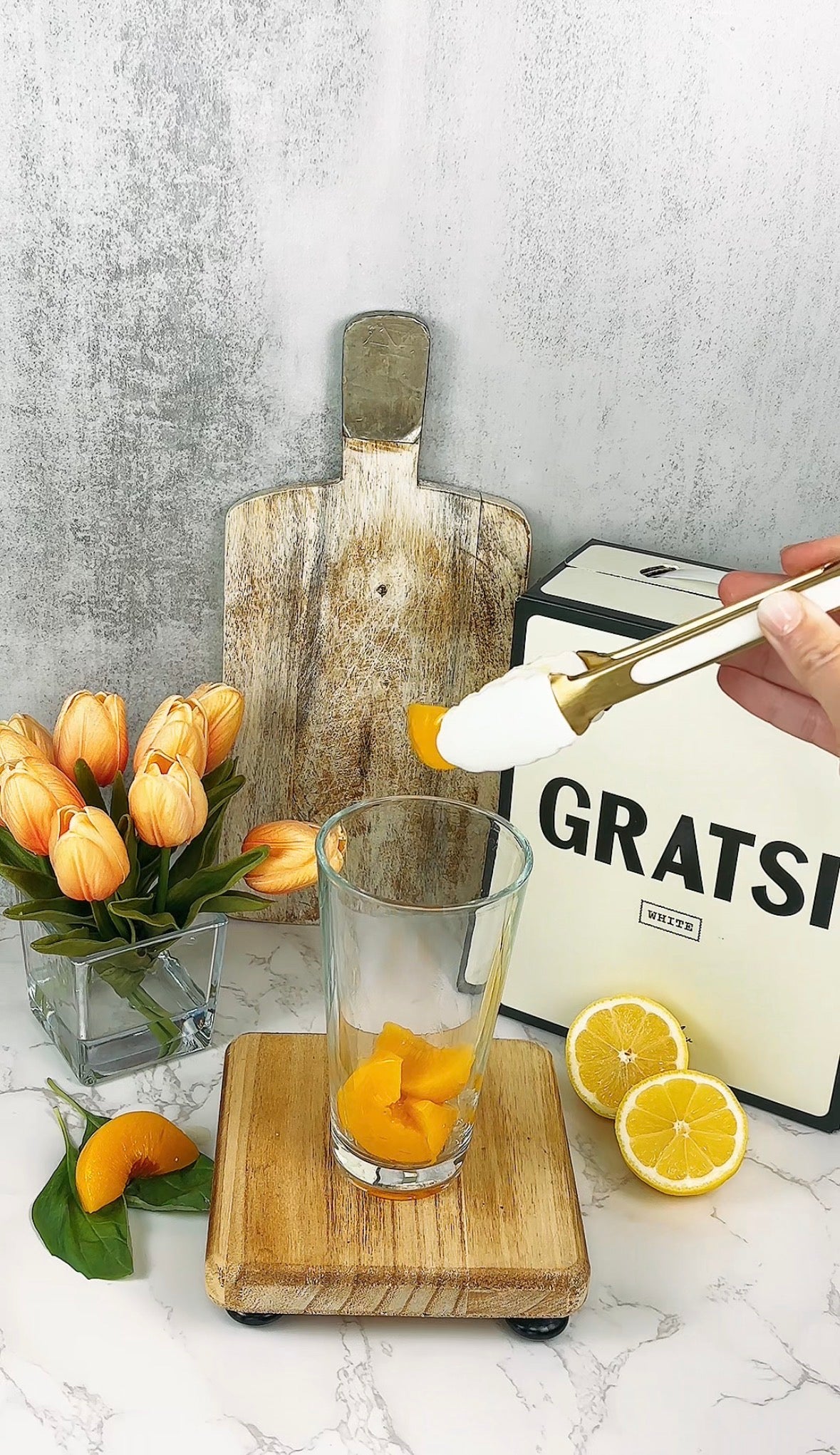 A hand using tongs places ice into a glass with orange slices. The glass is on a wooden board beside a halved lemon, a vase of orange tulips, a rustic cutting board, and a GRATSY box.