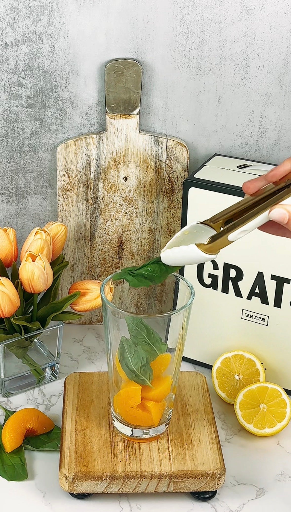 A hand uses tongs to place a green basil leaf into a glass containing orange fruit pieces. The glass sits on a wooden board, with cut lemons, a vase of orange tulips, and a rustic cutting board in the background.