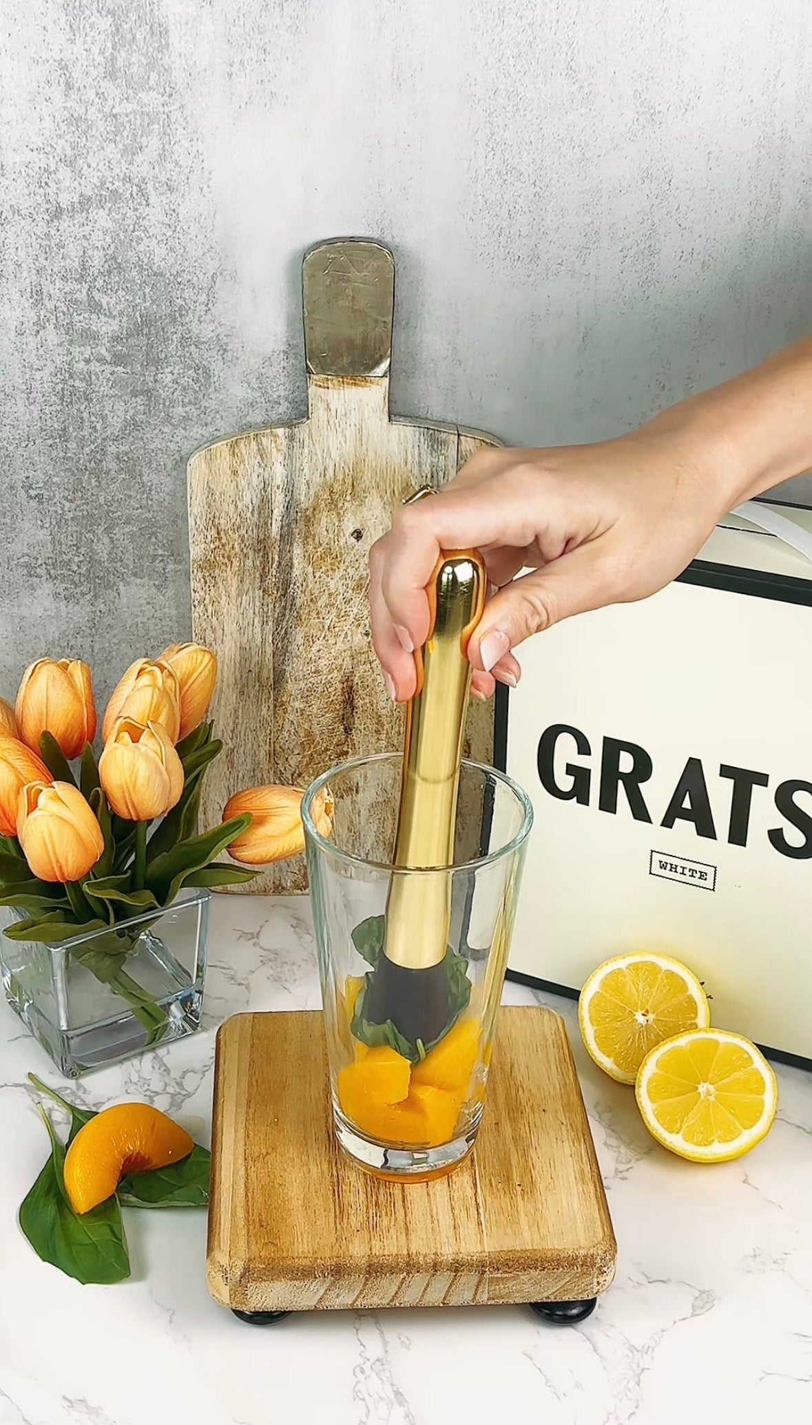 A hand uses a gold muddler to crush orange slices and mint leaves in a glass on a wooden board. Nearby are tulips in a vase, a halved lemon, and a box with the word GRATS on it.