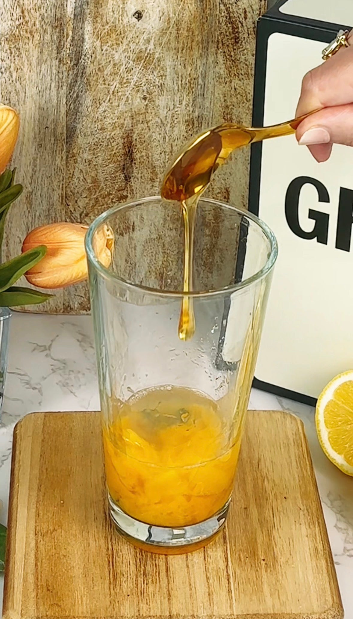 A hand drizzles honey from a spoon into a glass containing a yellow mixture, possibly egg yolk. The glass sits on a wooden board beside a sliced lemon and flowers, with a partially visible sign in the background.