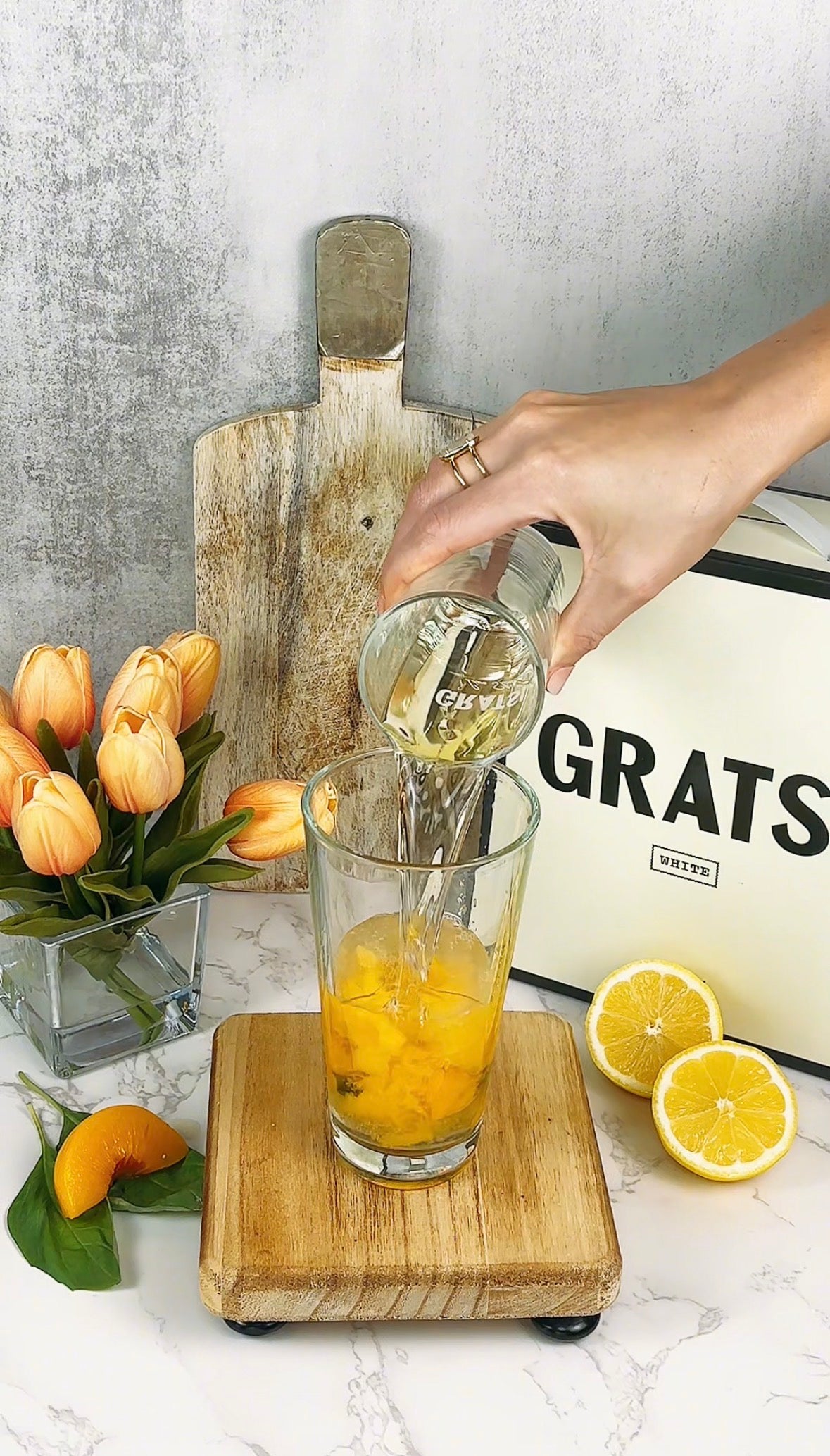 A hand pours a clear liquid from a glass into a tall glass containing orange pieces. The setup includes a cutting board, a bouquet of orange tulips, halved lemons, and a sliced orange on a marble countertop.