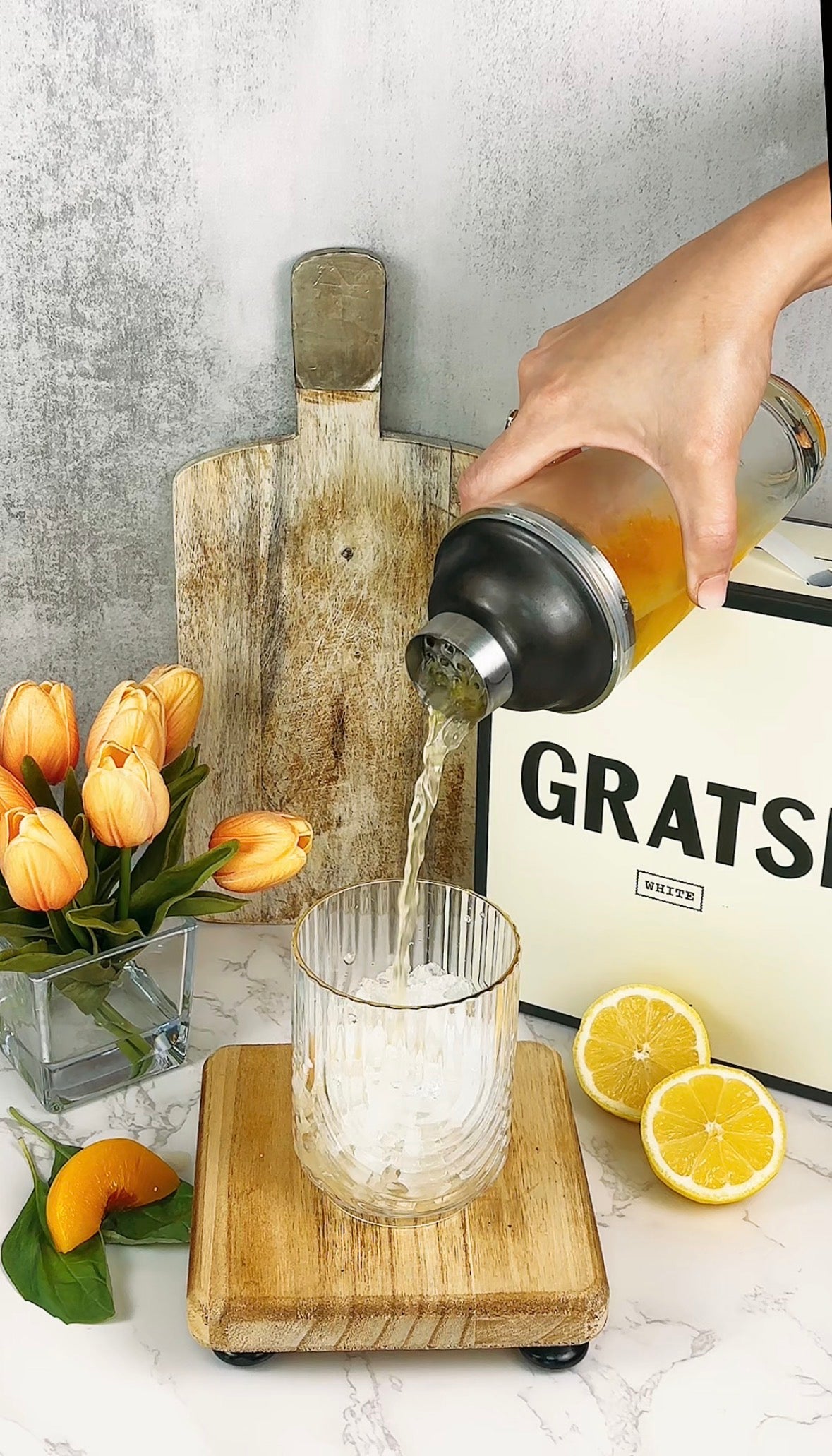 A hand pours a drink from a shaker into a glass with ice. Nearby are a cutting board, a vase of orange tulips, lemon halves, an orange slice, and a box labeled GRATS. The scene is set on a marble surface.