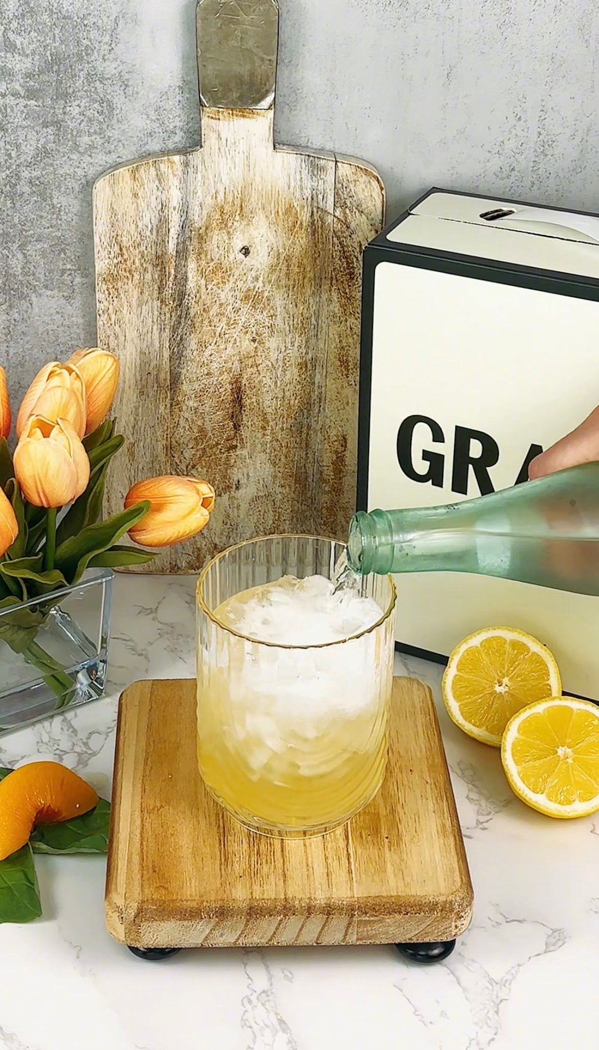 A hand pours sparkling water from a bottle into a glass with ice and yellow liquid, surrounded by tulips, a sliced lemon, and a peach on a marble counter, with a wooden board and a box labeled GRAB in the background.