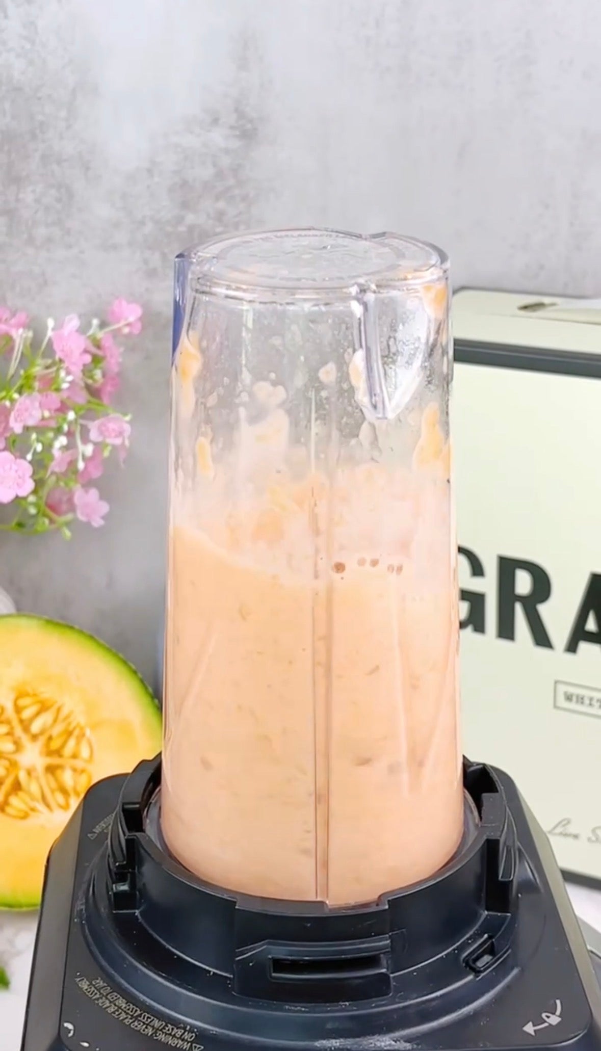 A blender cup filled with a creamy orange smoothie sits on its base. In the background, there are pink flowers, a pale green box labeled GRA, and a sliced melon with visible seeds.