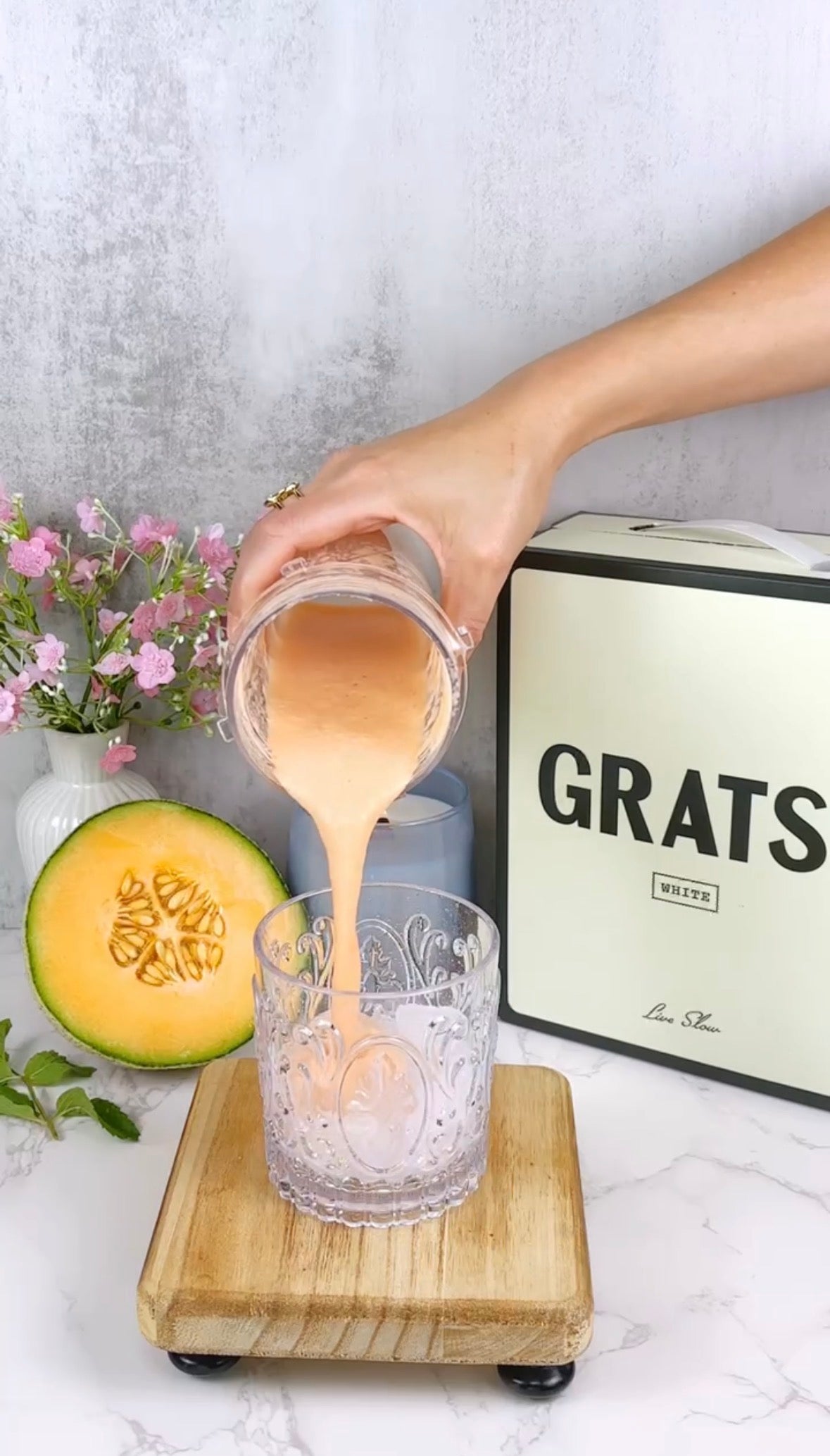 A hand pours a peach-colored smoothie from a glass into an ornate clear glass. Nearby are pink flowers, half a cantaloupe, a mint sprig, and a cream-colored box labeled GRATS. All items rest on a marble counter.