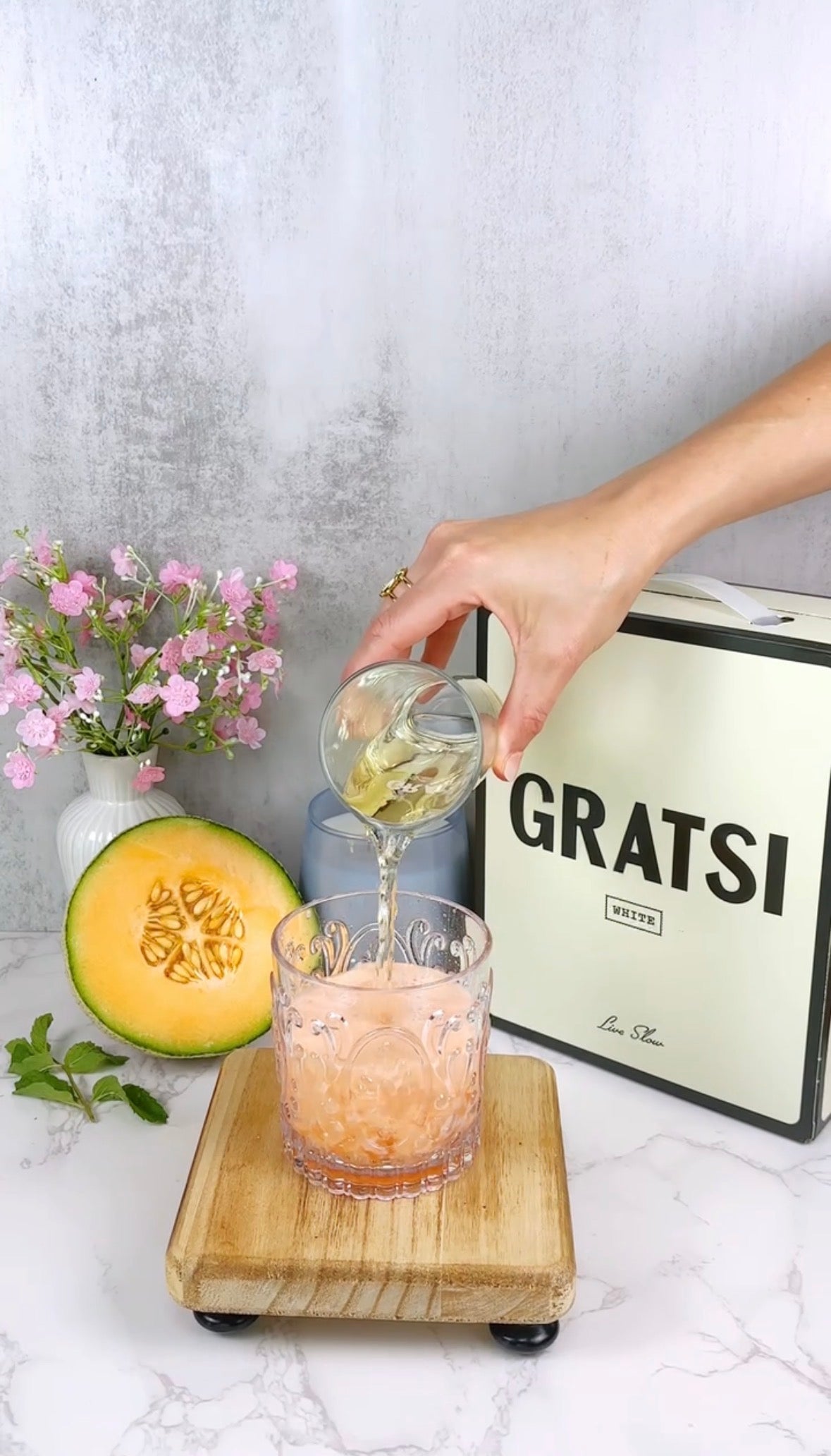 A hand pours a clear liquid into a glass with an orange drink. Nearby are a vase of pink flowers, a half cantaloupe, mint leaves, a GRATS! box, and a glass pitcher on a wooden board against a gray background.