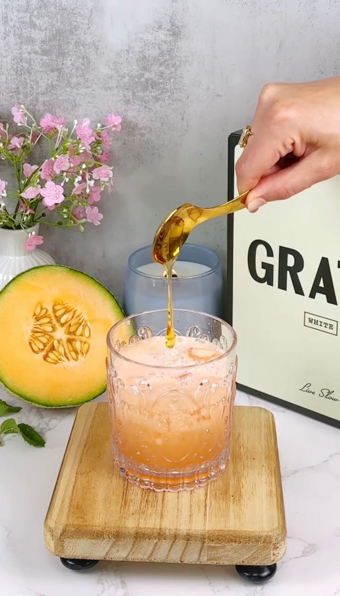A hand drizzles honey from a golden spoon into a glass of melon-colored drink on a wooden board. A half cantaloupe, pink flowers, and a framed sign are in the background.