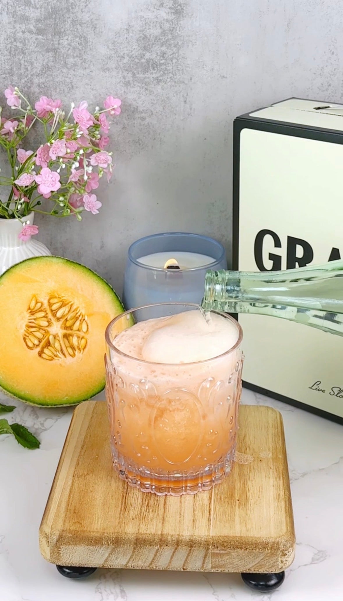 A glass of orange melon drink with foam on top is being poured from a bottle. Beside it are a halved cantaloupe, a candle, a flower vase with pink blossoms, and a box labeled GRATEFUL.