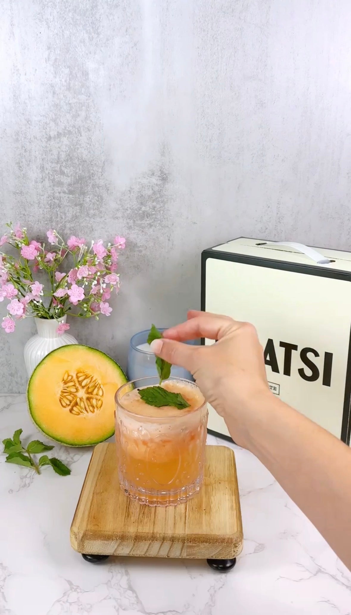 A hand garnishes a glass of orange melon drink with a mint leaf. Beside the drink are a halved melon, sprigs of mint, a vase of pink flowers, and a white box labeled SATSI on a light surface.