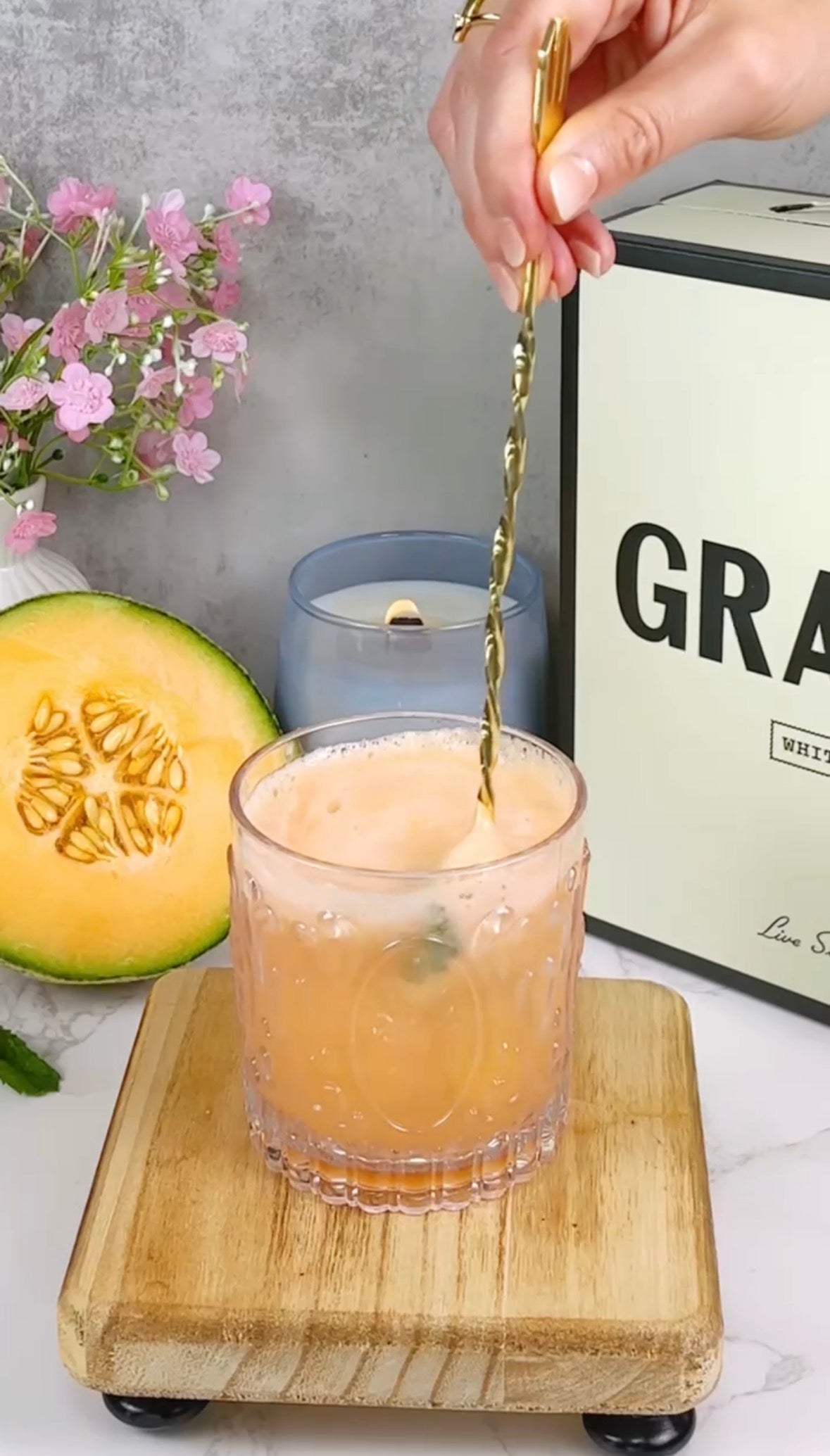 A hand stirs an orange cocktail in a glass with a golden twisted spoon. The drink is on a wooden board, next to a candle, a half cantaloupe, pink flowers, and a box labeled GRAS.