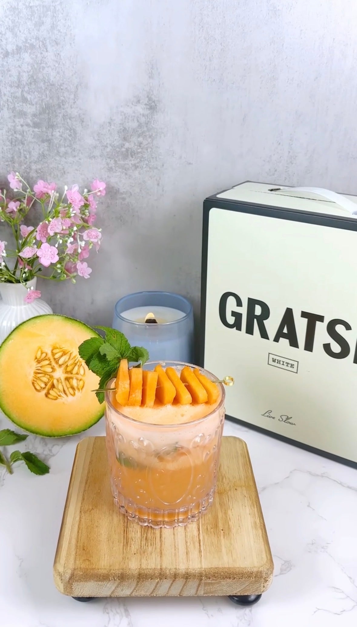 A glass of orange melon cocktail garnished with melon slices and mint sits on a wooden board. Beside it are a halved cantaloupe, a candle, pink flowers in a vase, and a box labeled GRATS.