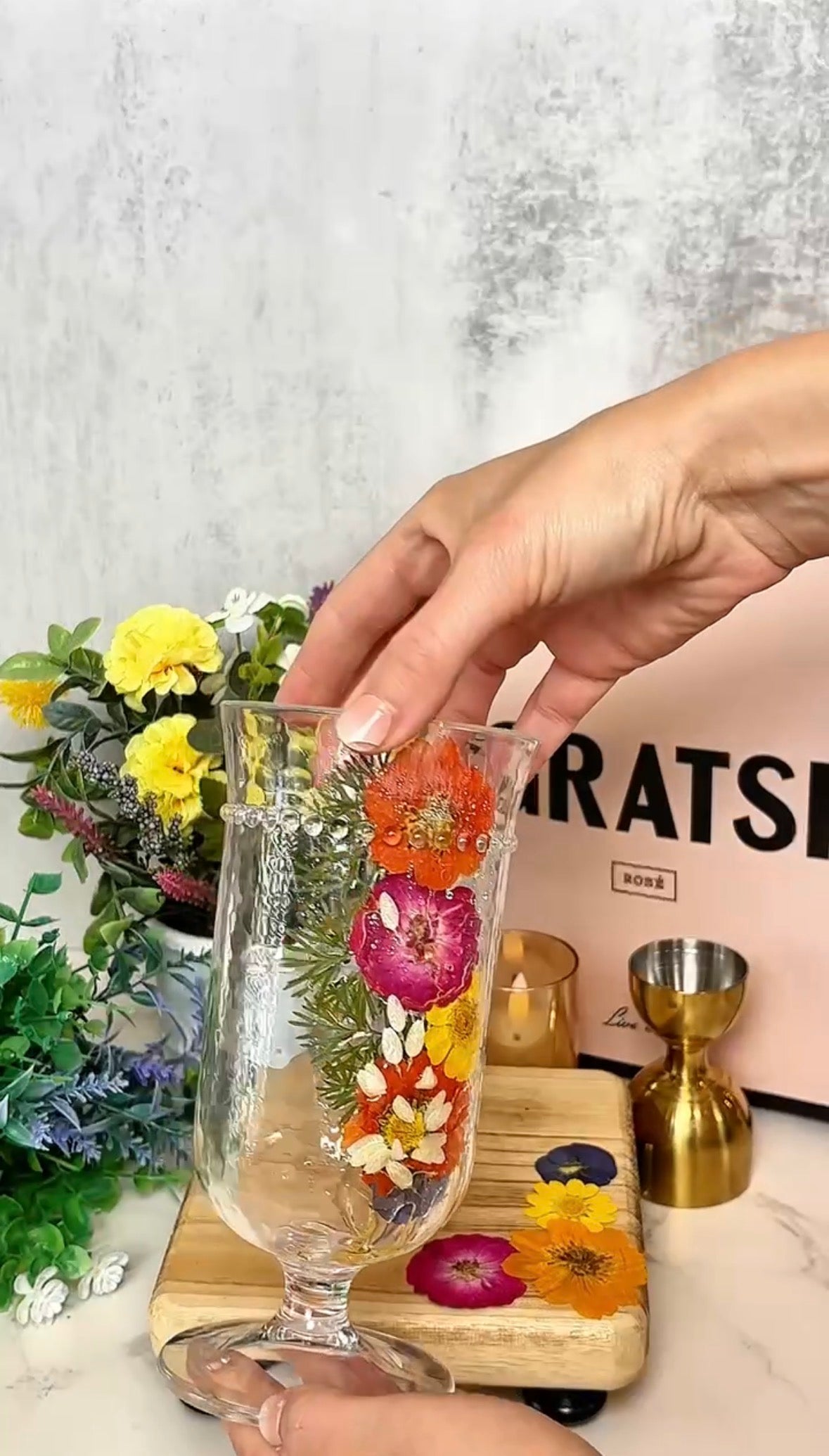 A hand places a clear glass decorated with colorful pressed flowers on a wooden board. In the background are fresh flowers, a pink box, and gold cocktail tools.