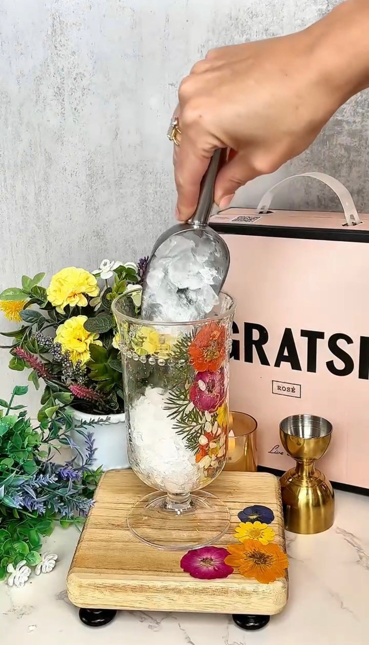 A hand scoops ice into a tall glass decorated with colorful pressed flowers. The glass sits on a wooden board with more flowers, surrounded by bar tools and a pink box labeled GRATSY, with floral arrangements in the background.