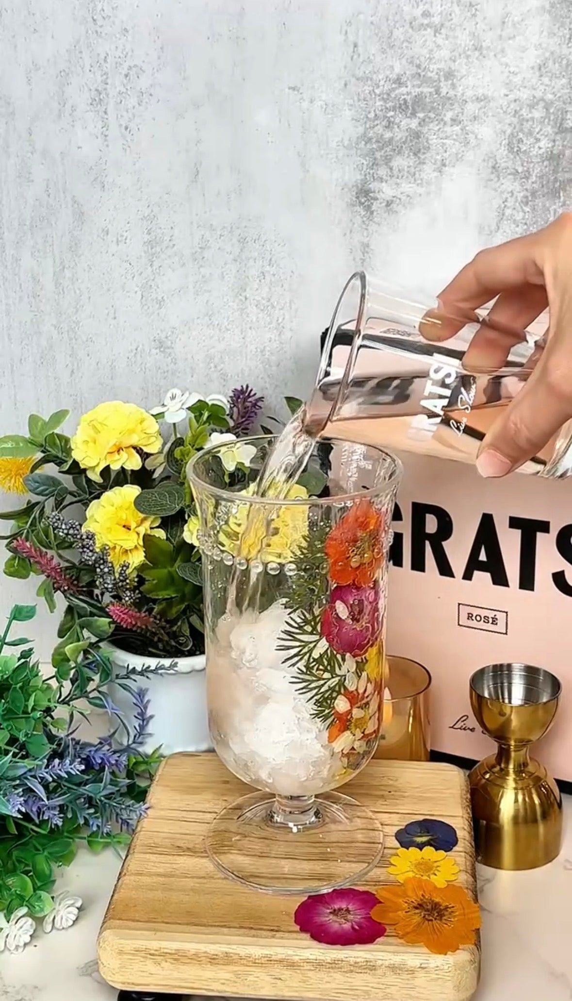 A hand pours a clear liquid into a floral glass filled with ice, beside a wooden board with measuring cups, a bouquet of yellow flowers, and a pink box labeled GRATS ROSÉ.