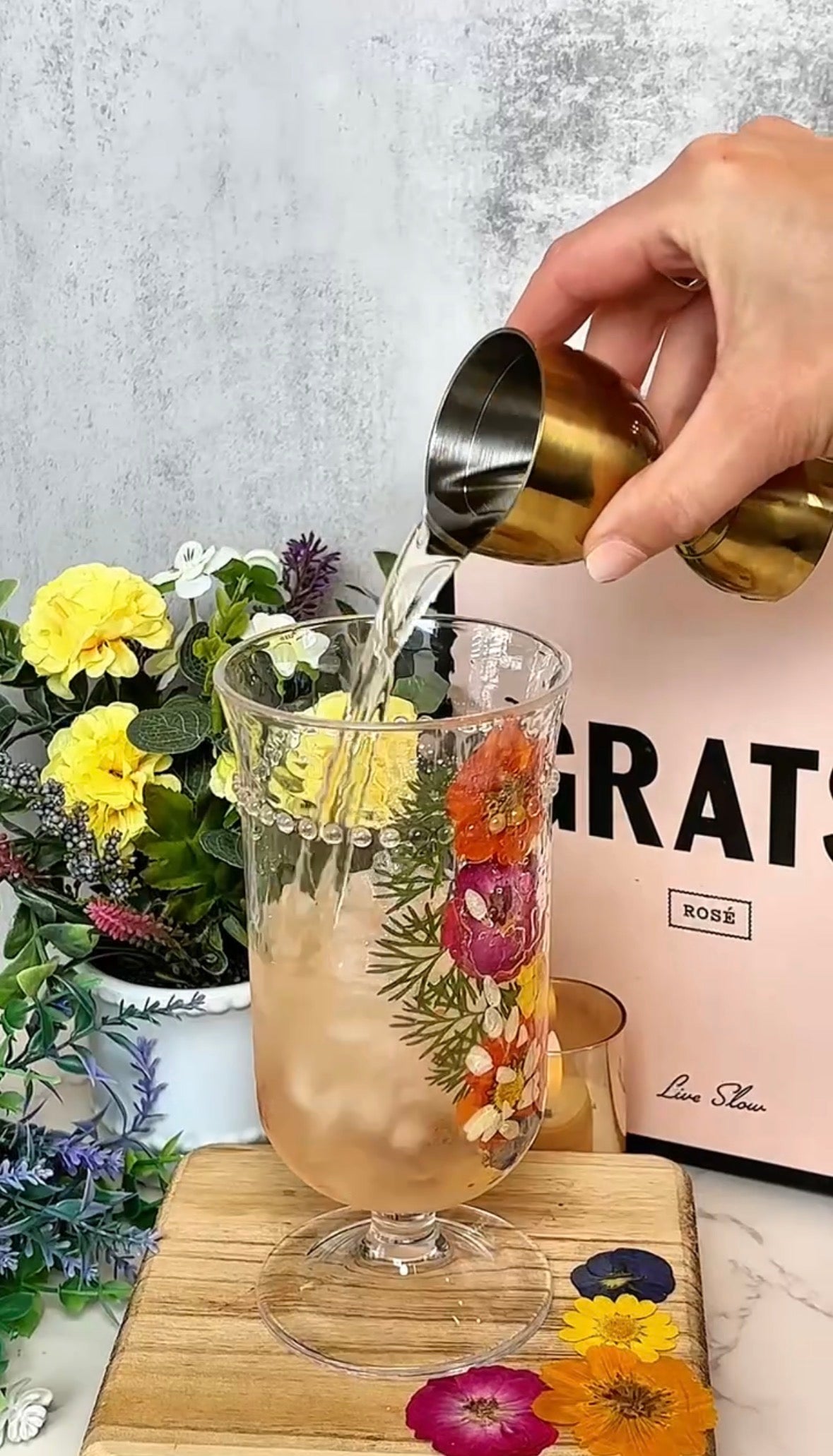 A hand pours clear liquid from a gold jigger into a floral glass filled with ice. The glass sits beside a bouquet of yellow and purple flowers and a pink box with GRATS written on it.