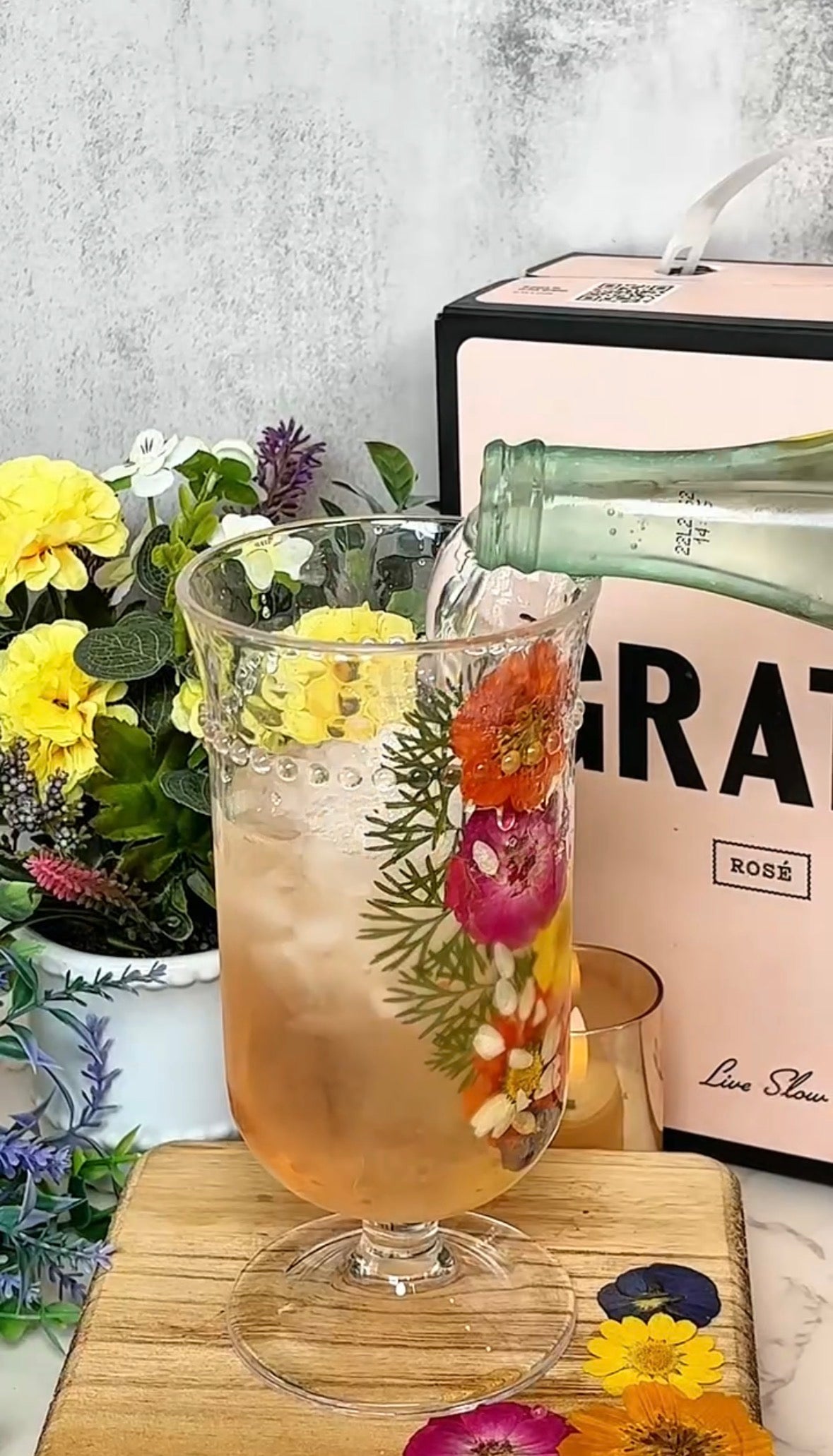A glass with ice and colorful edible flowers is being filled with sparkling wine. In the background, there are yellow flowers and a pink box labeled ROSÉ. The setting appears bright and decorative.