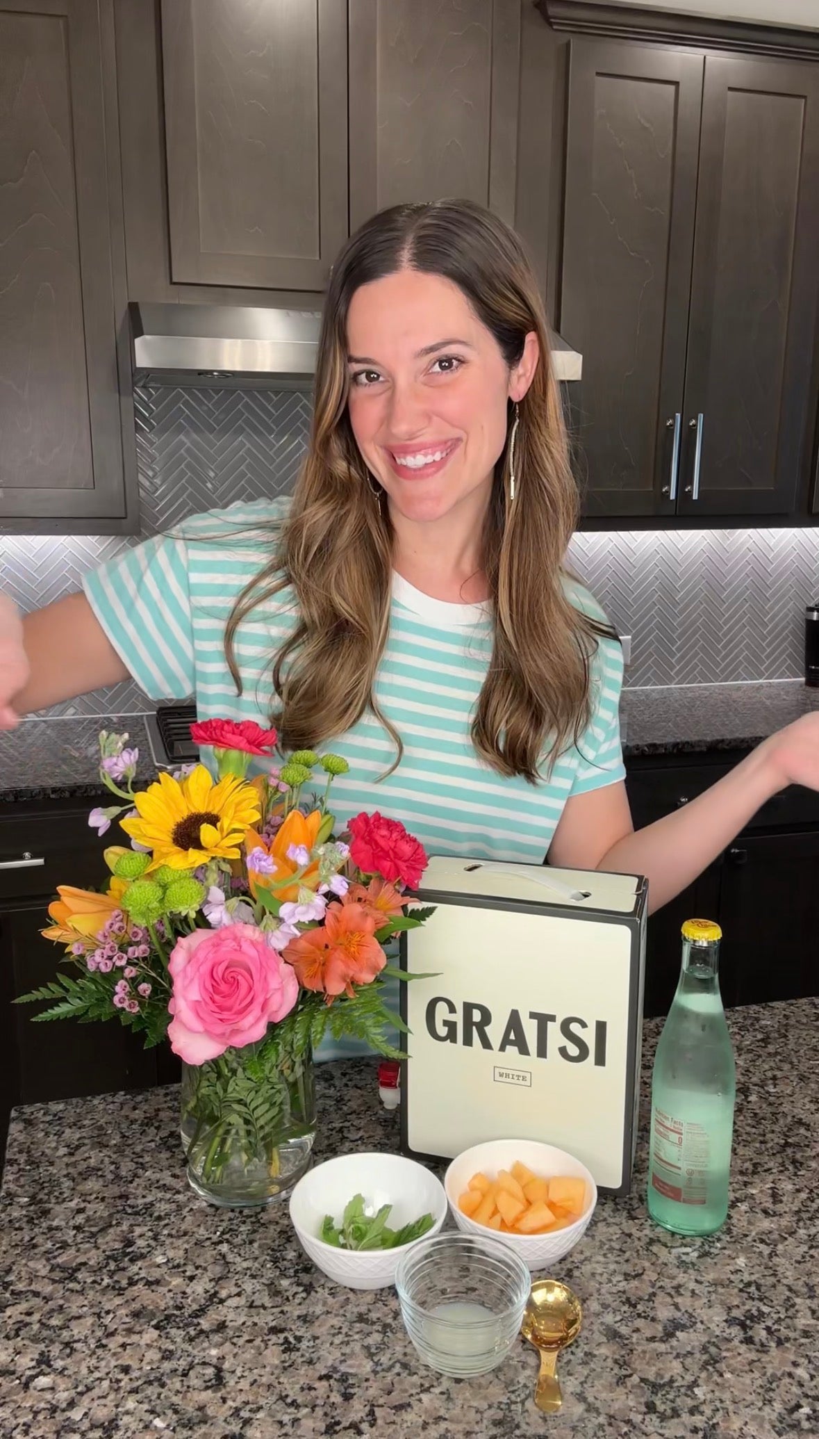 A smiling woman in a striped shirt stands in a kitchen with a Gratsi box, a bouquet of colorful flowers, a bottle of sparkling water, a bowl of melon, mint leaves, and a jigger on the counter.