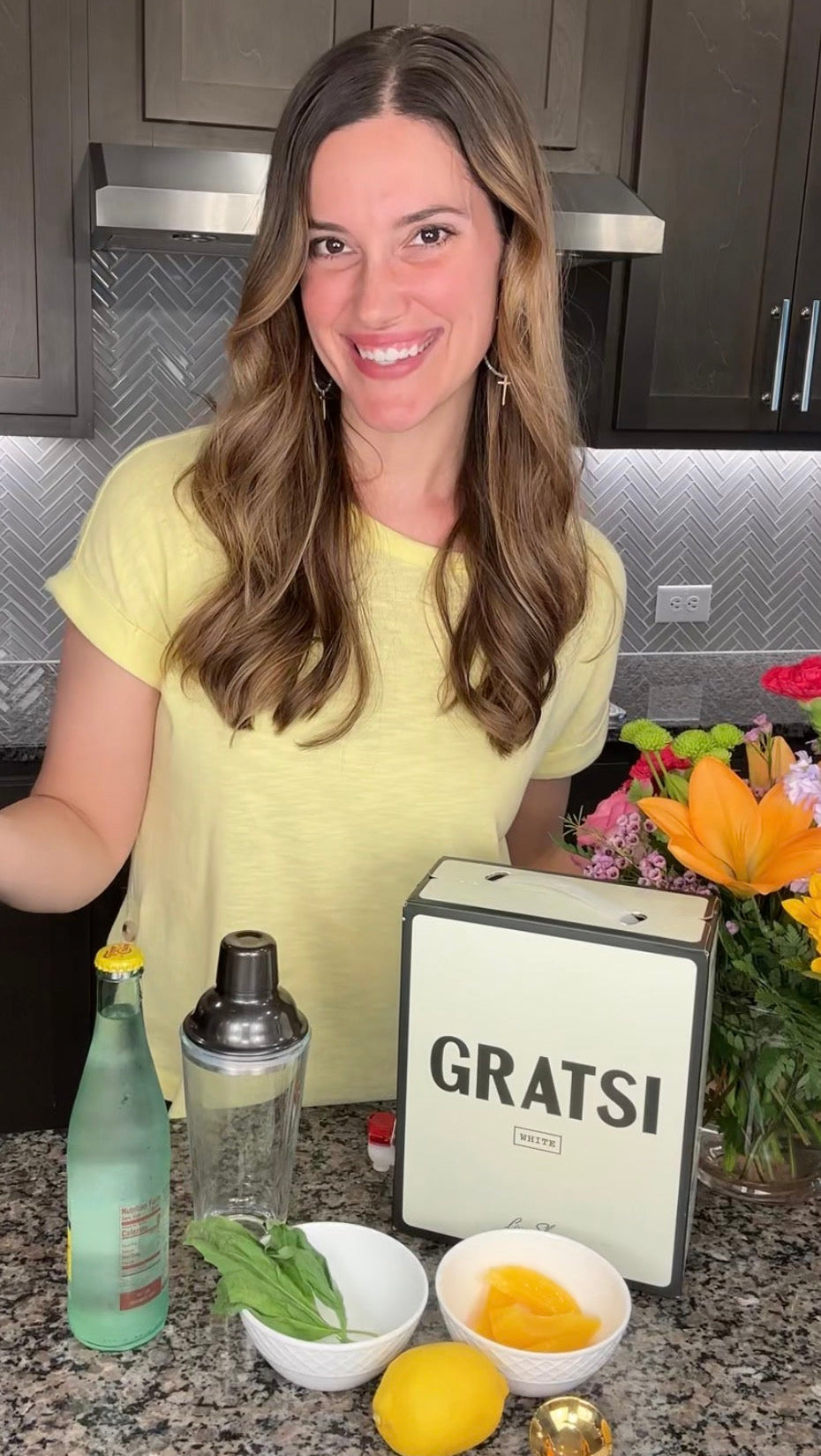 A woman with long brown hair wearing a yellow shirt stands in a kitchen, smiling. In front of her are a cocktail shaker, a bottle, a GRATS! box, a bouquet of flowers, lemon, basil, and a small bowl with yellow pieces.