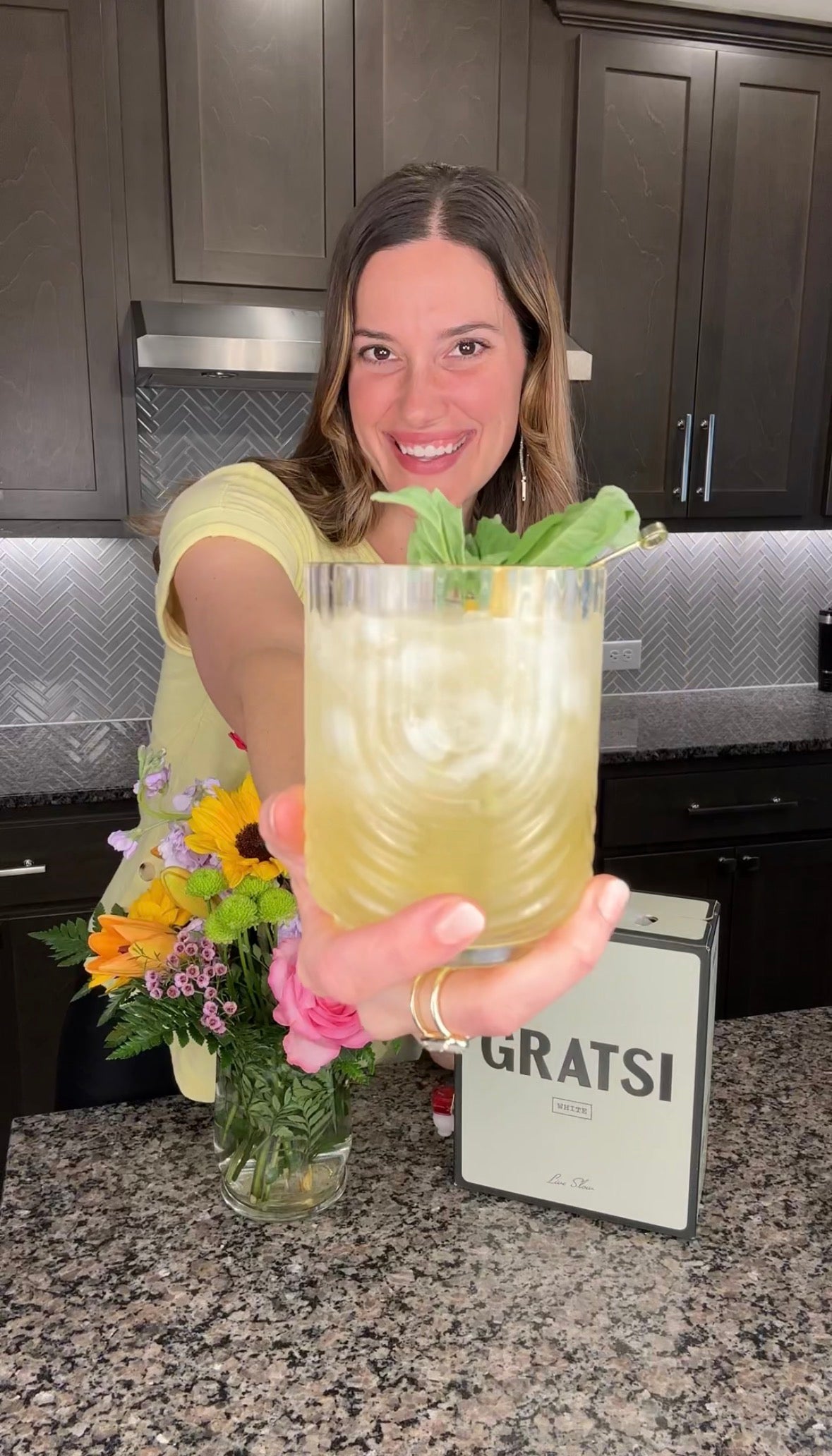 A woman in a yellow shirt stands in a kitchen, smiling and holding a cocktail garnished with a basil leaf toward the camera. A flower bouquet and a box labeled “GRATSI” are on the counter.